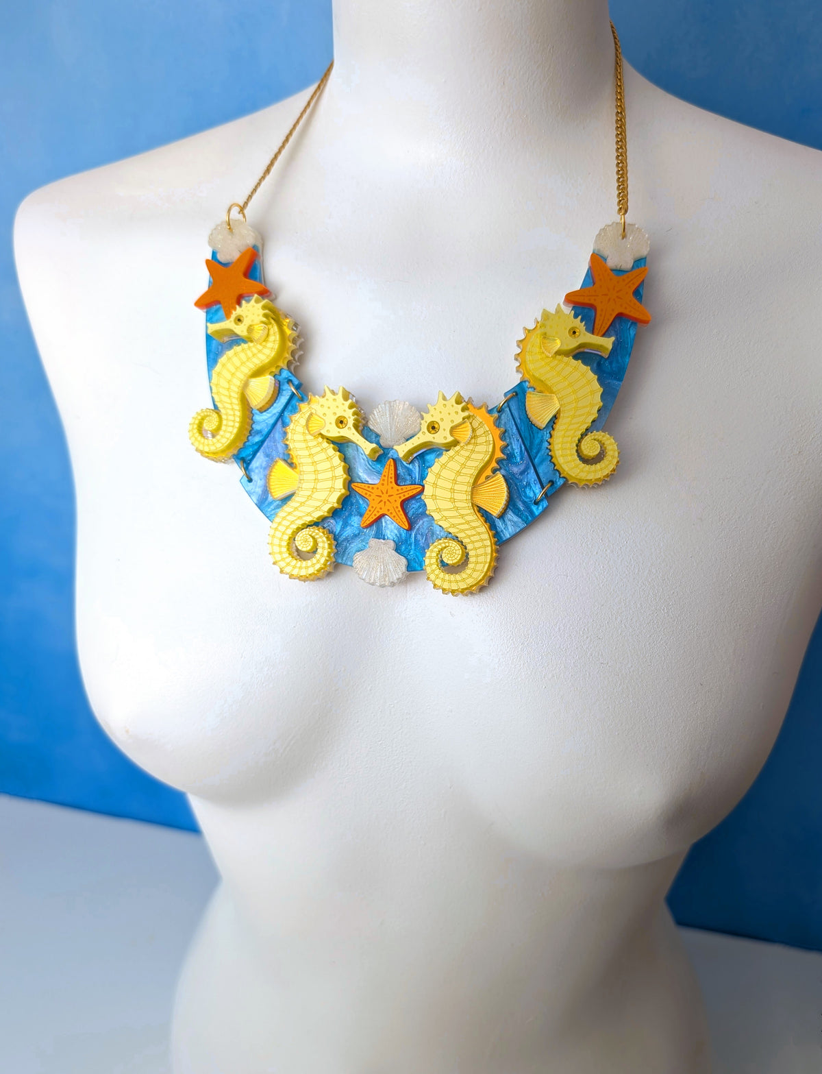 A handmade seahorse-themed statement necklace displayed on a white mannequin bust. The necklace features yellow seahorses, orange starfish, and a blue and white patterned base with gold-toned chain.