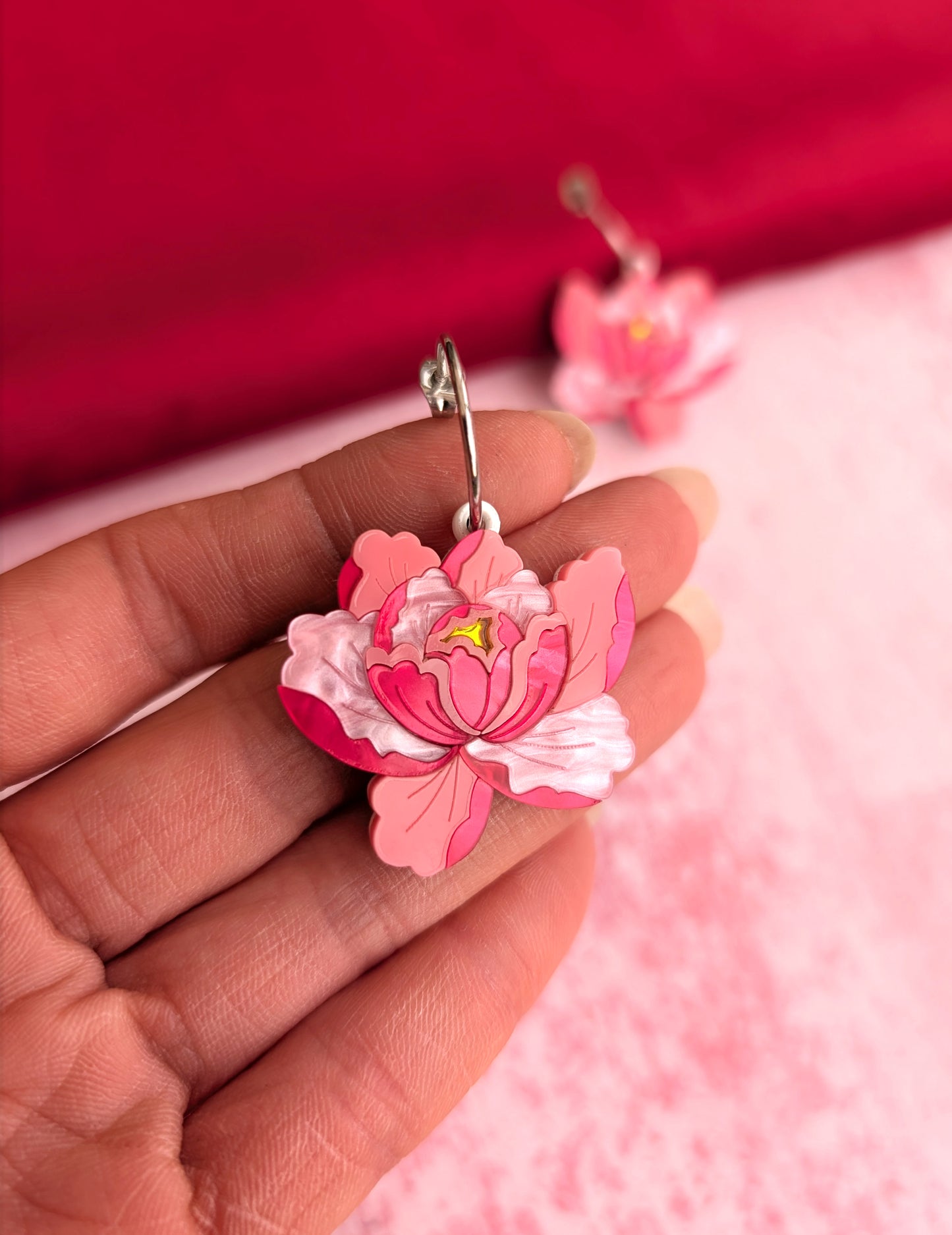 A pair of pink floral design earrings, resembling peony flowers, attached to the ear with a stud. The earrings are held in a person's hand against a red and pink background.