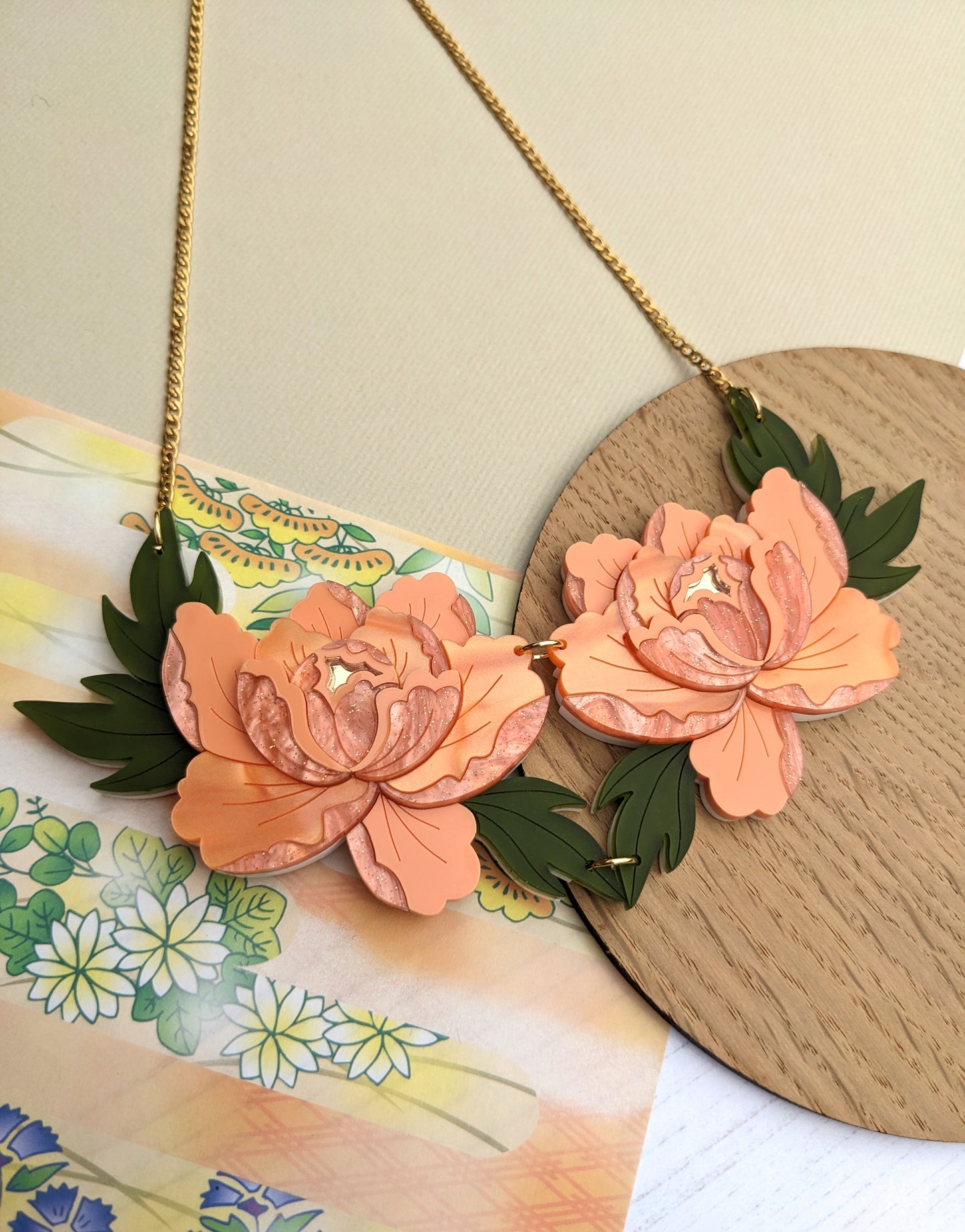 A double peony necklace with apricot and green acrylic flowers, set against a pearl marble and gold mirror acrylic background with a gold chain.