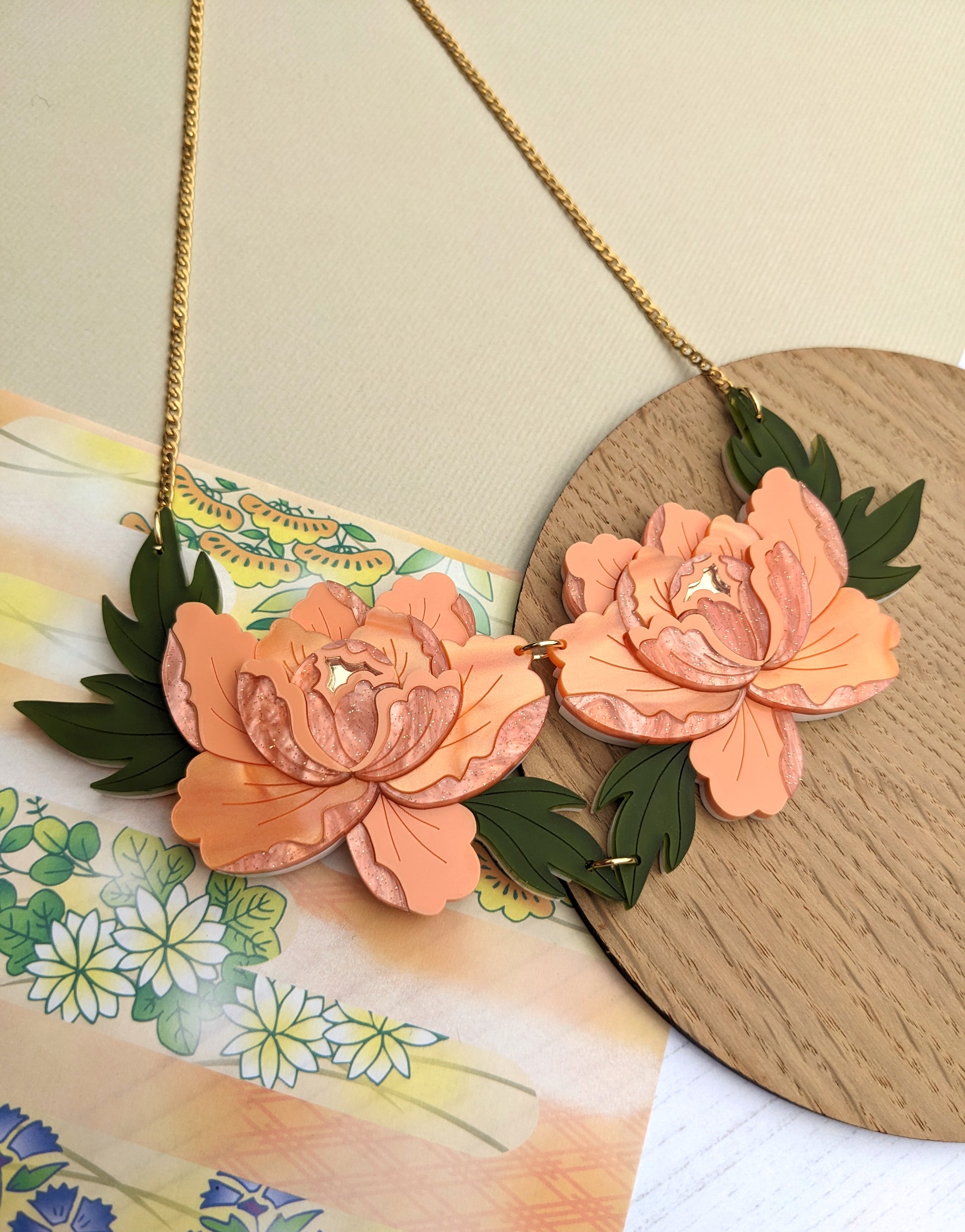 A double peony necklace with apricot and green acrylic flowers, set against a pearl marble and gold mirror acrylic background with a gold chain.