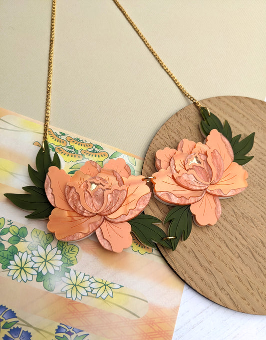 A double peony necklace with apricot and green acrylic flowers, set against a pearl marble and gold mirror acrylic background with a gold chain.