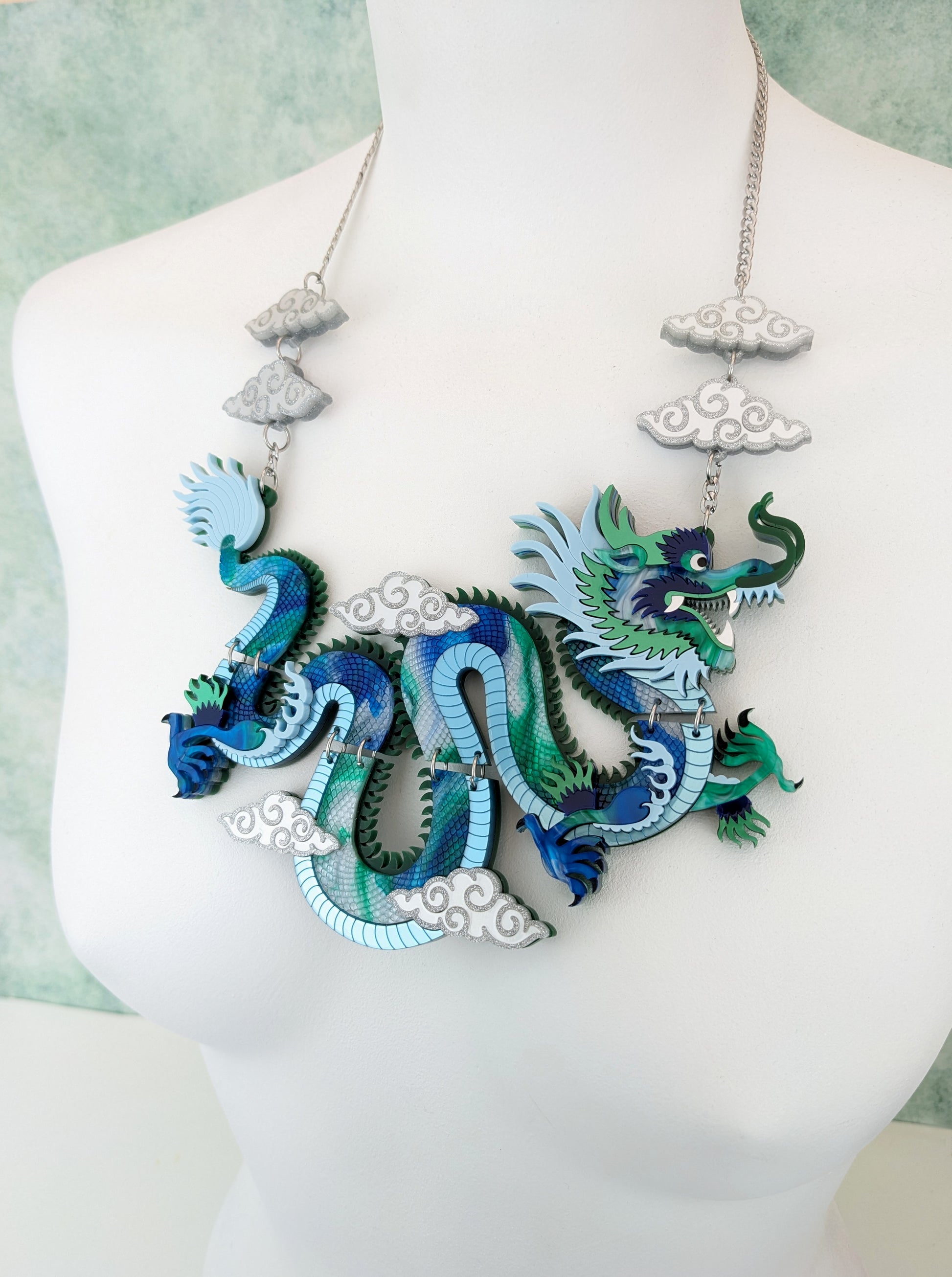 A statement necklace featuring a multicolored Chinese dragon design with blue, green, and white pearlescent marble acrylic, accompanied by cloud shapes, displayed on a mannequin.