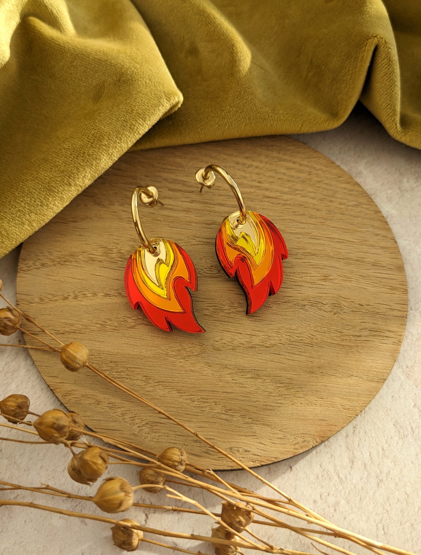 A pair of gold-colored hoop earrings with red, orange, and yellow flame-shaped acrylic charms, displayed on a wooden round tray with dried flowers and a green velvet fabric in the background.