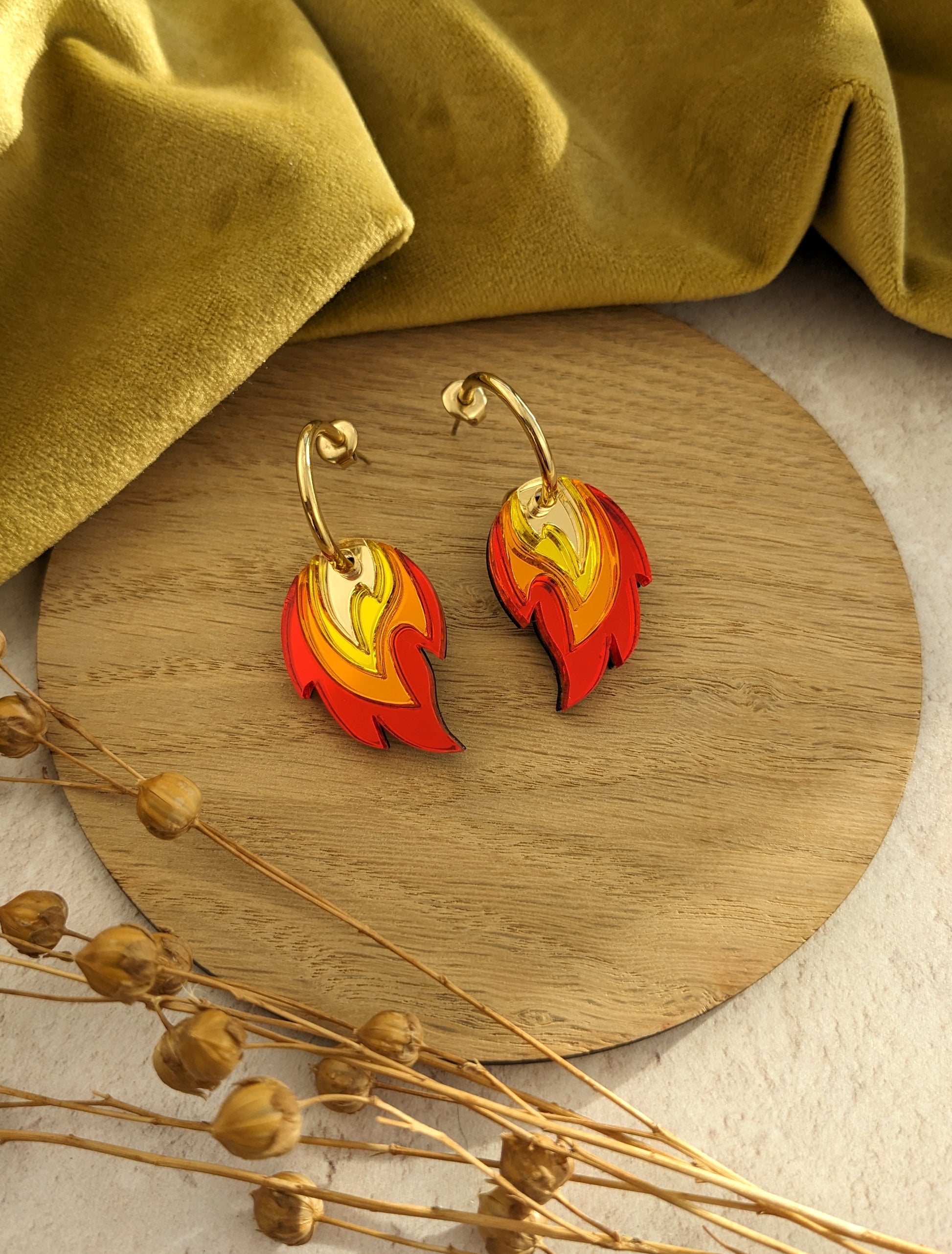A pair of gold-colored hoop earrings with red, orange, and yellow flame-shaped acrylic charms, displayed on a wooden round tray with dried flowers and a green velvet fabric in the background.