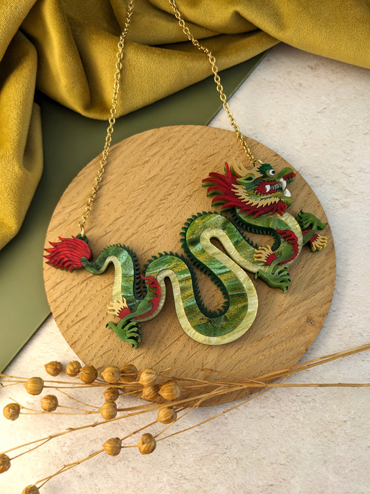 A small, colorful acrylic necklace designed in the shape of a Chinese dragon, displayed against a wooden background with yellow fabric and decorative wheat.