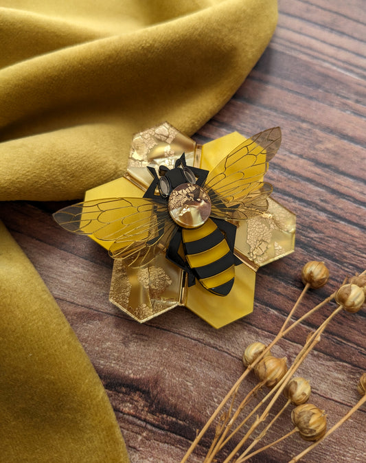A brooch depicting a honey bee on a honeycomb base, made with recycled acrylic in various shades of yellow, black, and transparent wings, placed on a wooden surface next to a yellow fabric.