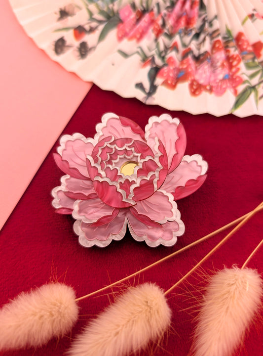 A brooch or pendant with a peony-inspired design in pale pink, white pearl marble acrylic, and gold mirror centre, displayed on a red background with floral patterns and light pink embellishments.