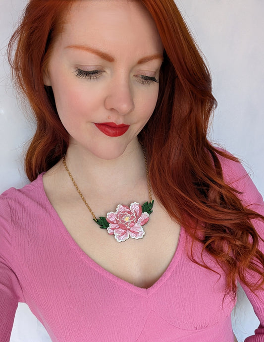 A person with red hair wearing a pink top and a floral necklace with gold-colored leaves.