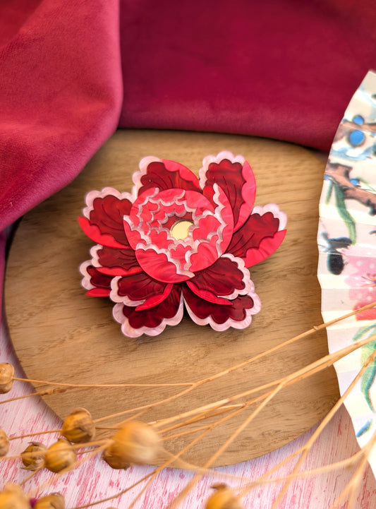 A red and pink floral brooch with a gold center, placed on a wooden surface with a patterned fabric edge.