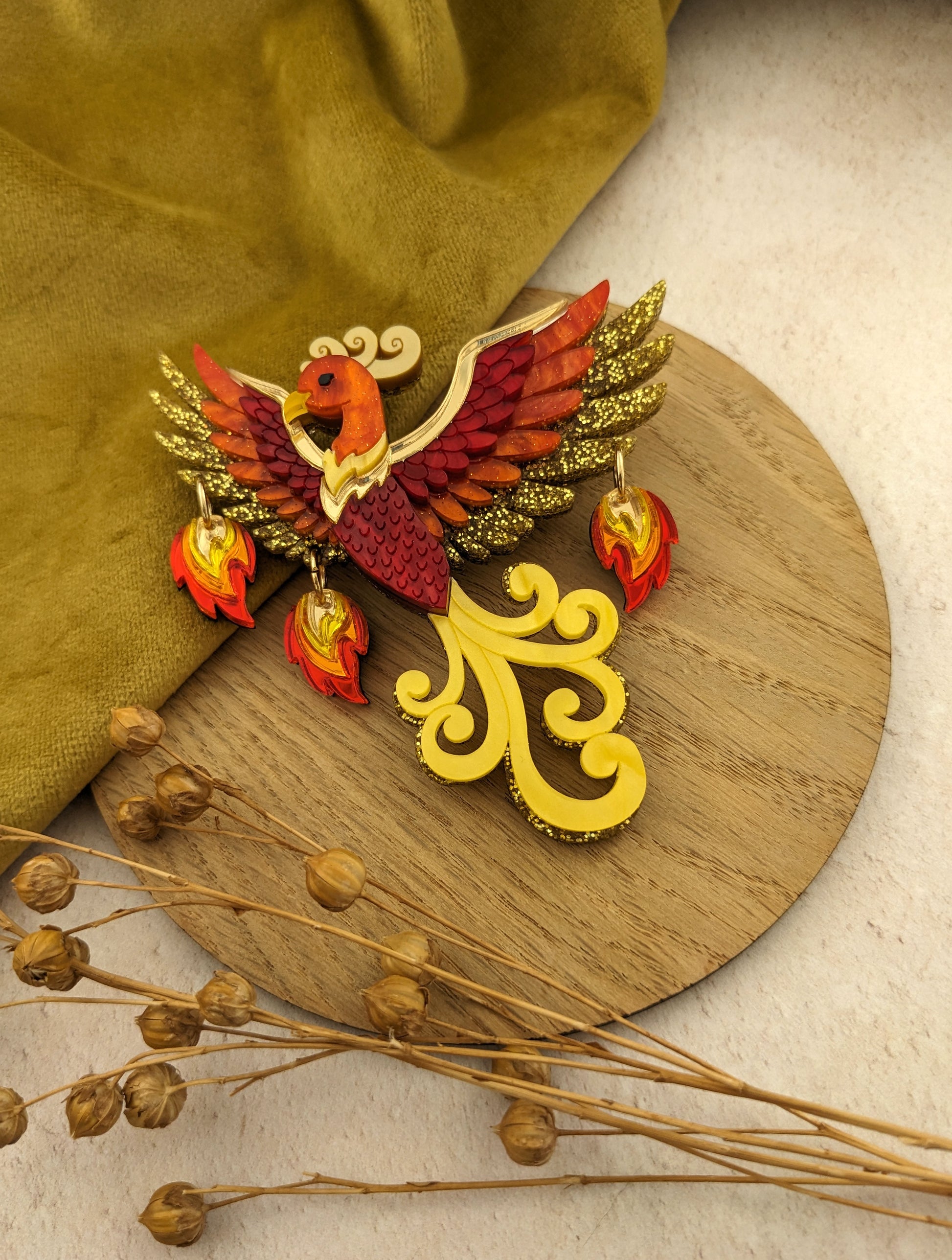 A brooch designed as a phoenix with flames, featuring yellow, red, and gold colors with glitter and pearl marble acrylic.