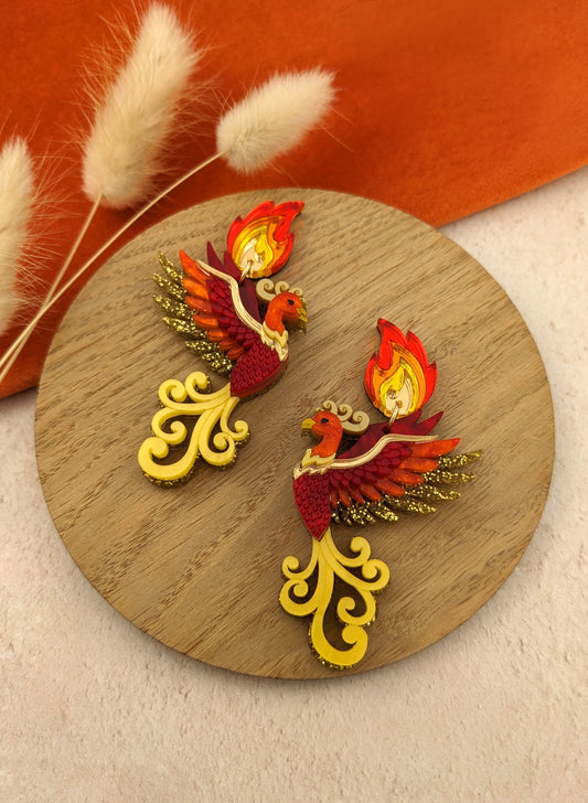 A pair of acrylic earrings designed to resemble a phoenix with flame details, featuring yellow, red, and orange colors against a wooden backdrop.
