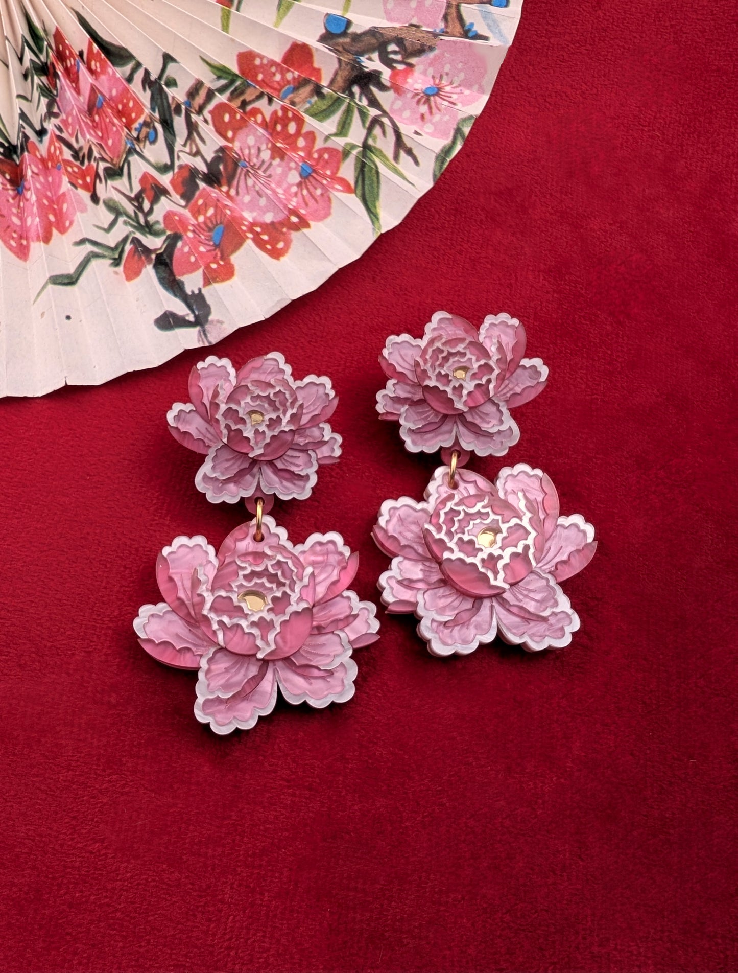 A pair of pale pink peony inspired statement earrings displayed on a red surface, with a floral patterned fabric in the background.