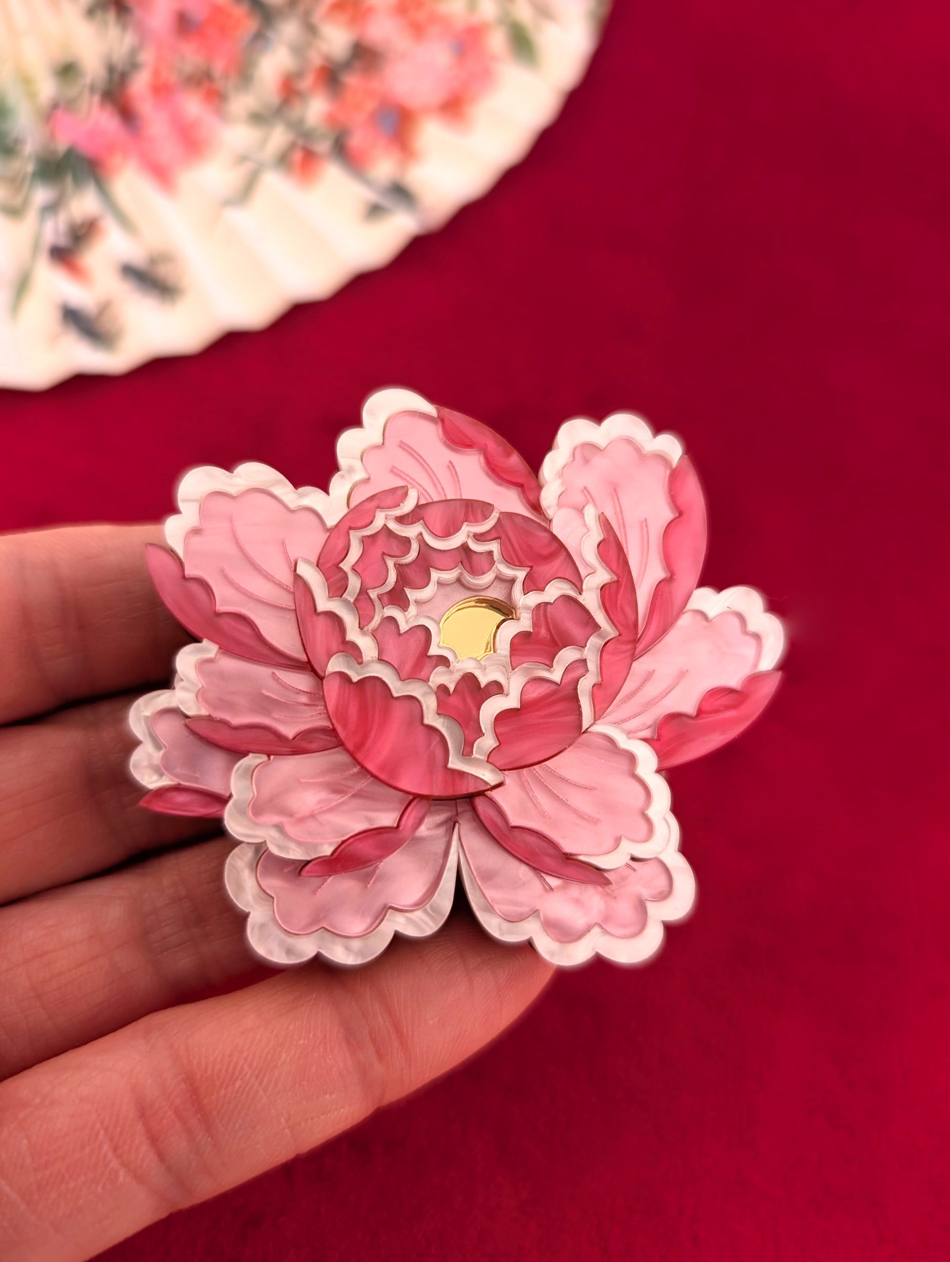 A brooch or pendant with a peony-inspired design in pale pink, white pearl marble acrylic, and gold mirror centre, displayed on a red background with floral patterns and light pink embellishments.