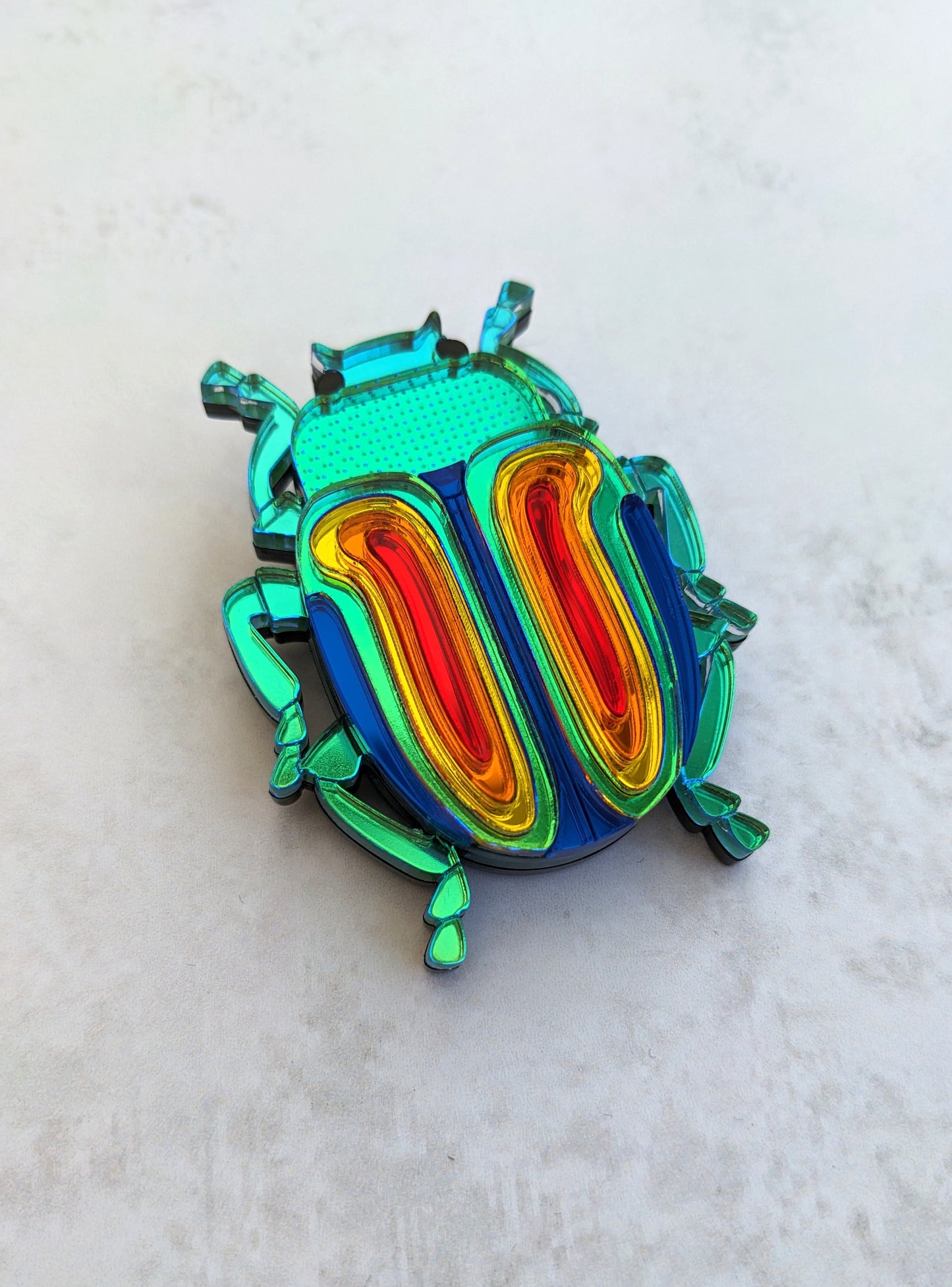 A colorful acrylic brooch designed in the shape of a rainbow beetle, with multiple layers of iridescent acrylic in shades of green, blue, red, and yellow.