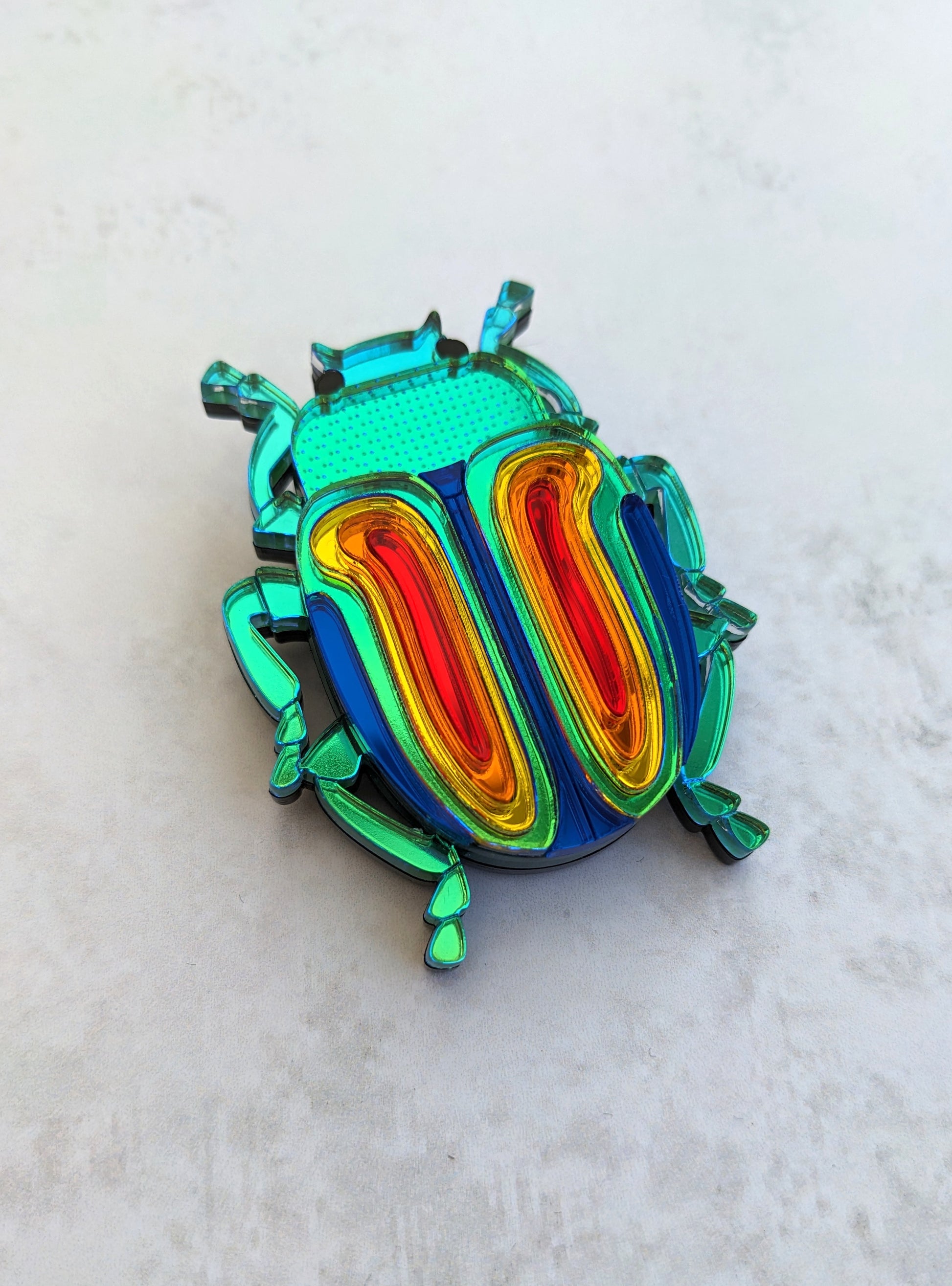 A colorful acrylic brooch designed in the shape of a rainbow beetle, with multiple layers of iridescent acrylic in shades of green, blue, red, and yellow.