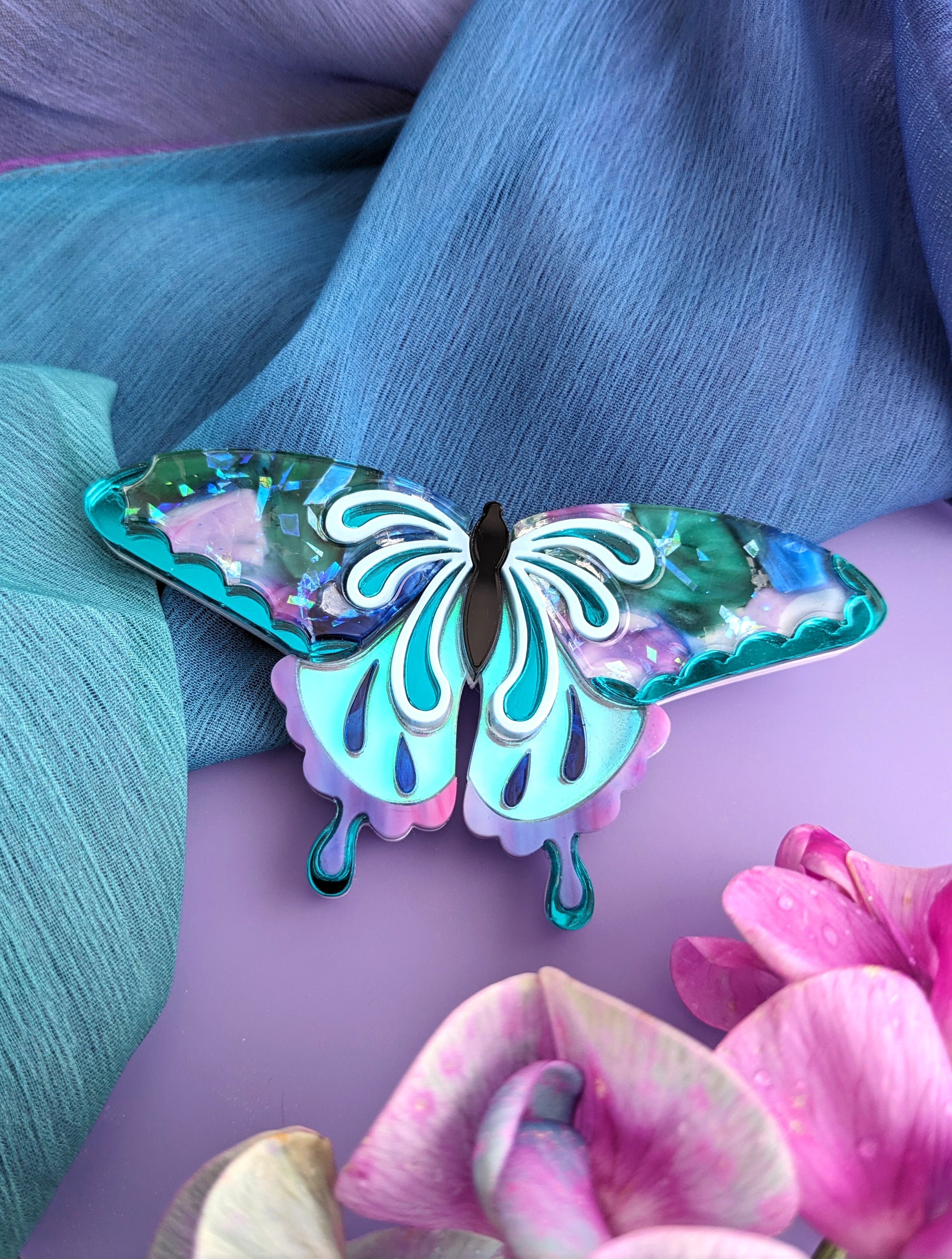 A brooch designed in the shape of a butterfly with wings featuring a mix of purple, teal, and blue colors, displayed against a purple and teal background with a pink flower in the lower right corner.
