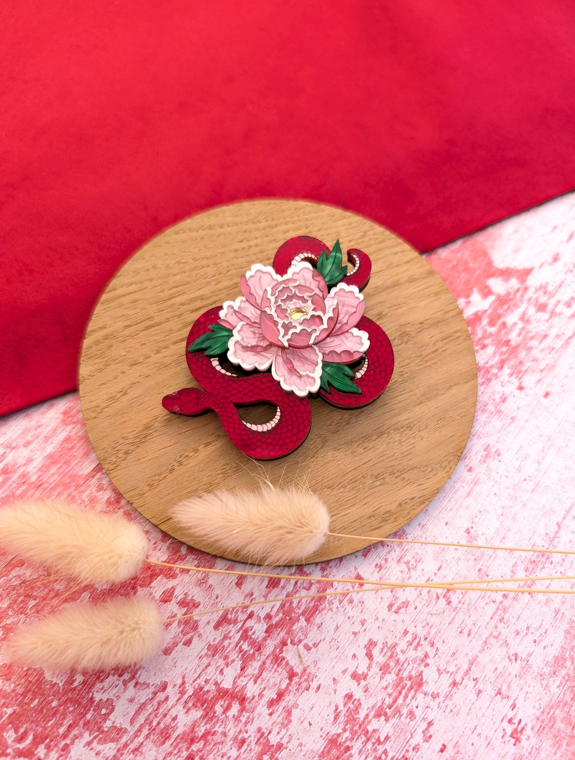 A red and gold brooch in the shape of a snake with a floral design, displayed on a wooden round stand with a red and white patterned background.