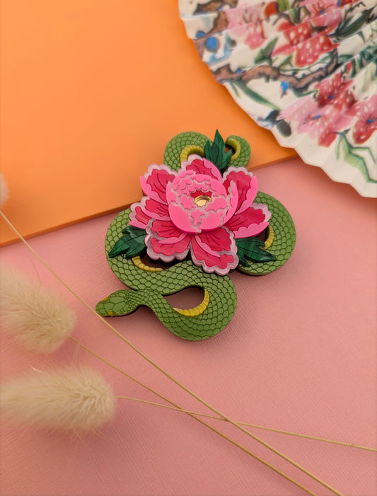 A green brooch designed with a snake and a floral motif, featuring a prominent pink flower, displayed against a pink and orange background with decorative elements.