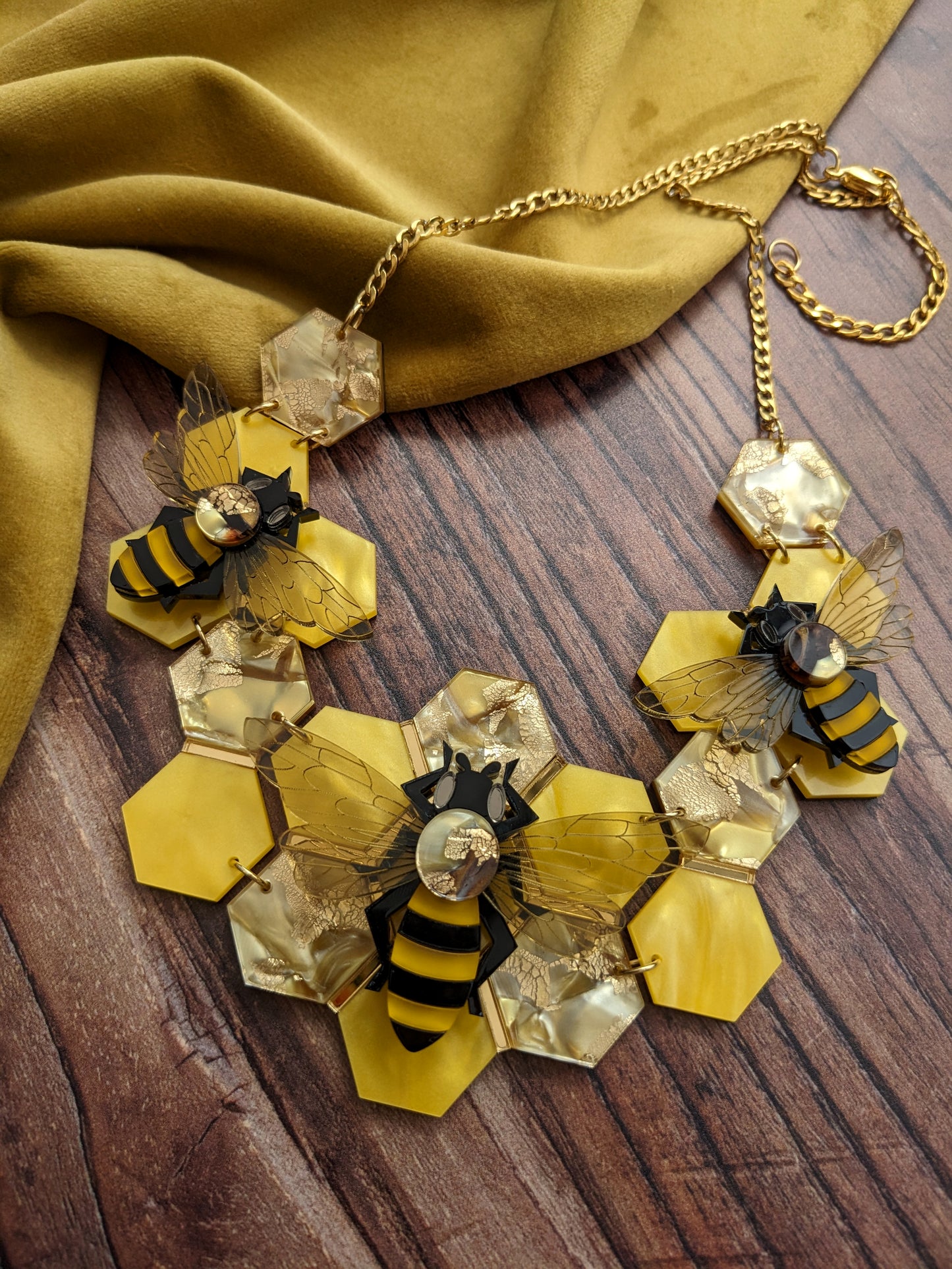 A statement honey bee necklace with a golden recycled acrylic honeycomb base and bees assembled on top, displayed on a wooden surface with a yellow fabric in the background.