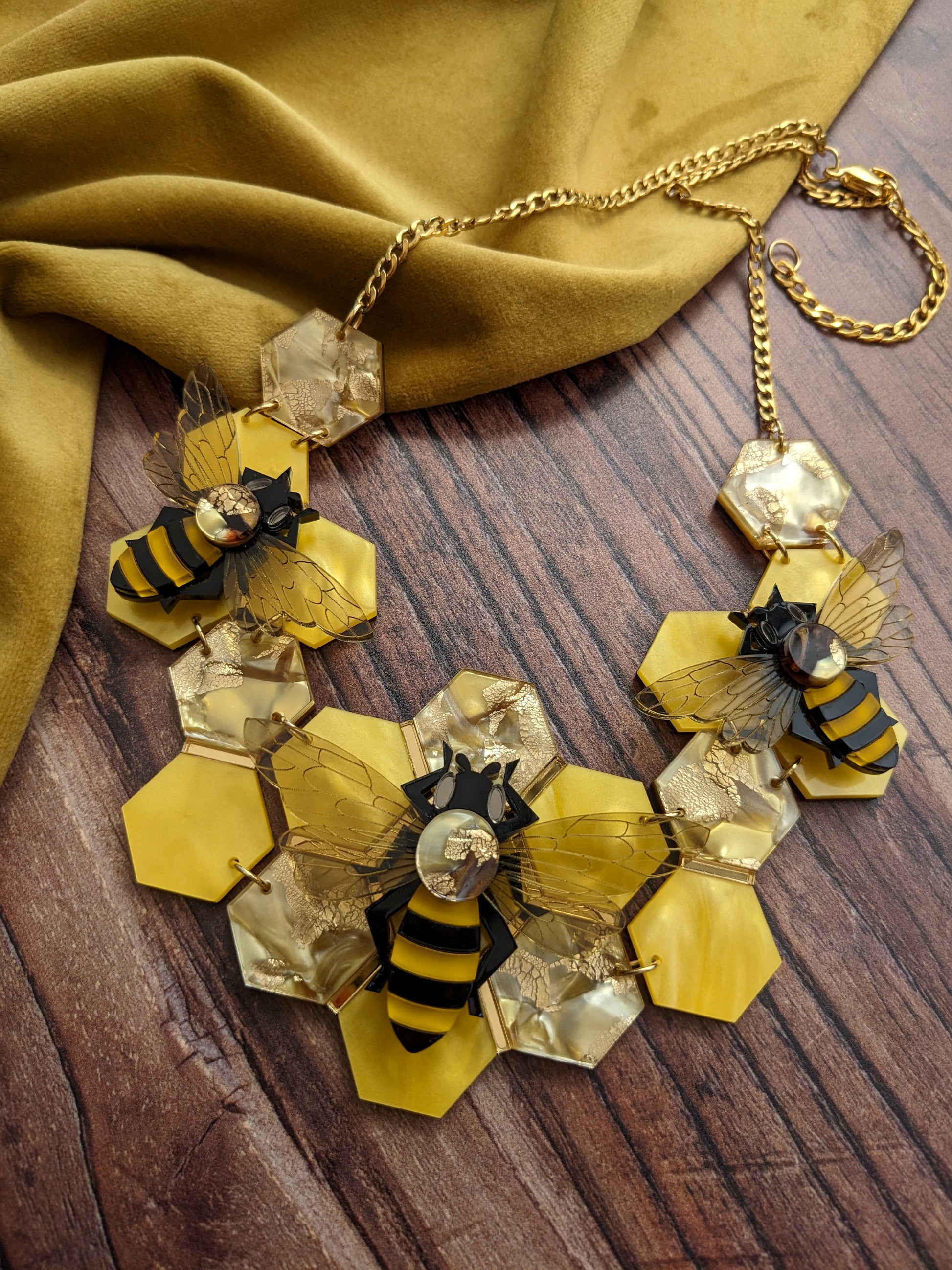 A statement honey bee necklace with a golden recycled acrylic honeycomb base and bees assembled on top, displayed on a wooden surface with a yellow fabric in the background.