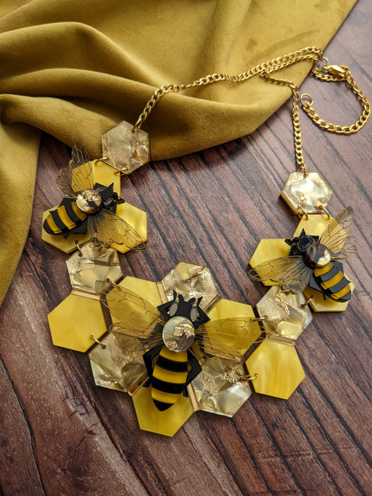 A statement honey bee necklace with a golden recycled acrylic honeycomb base and bees assembled on top, displayed on a wooden surface with a yellow fabric in the background.