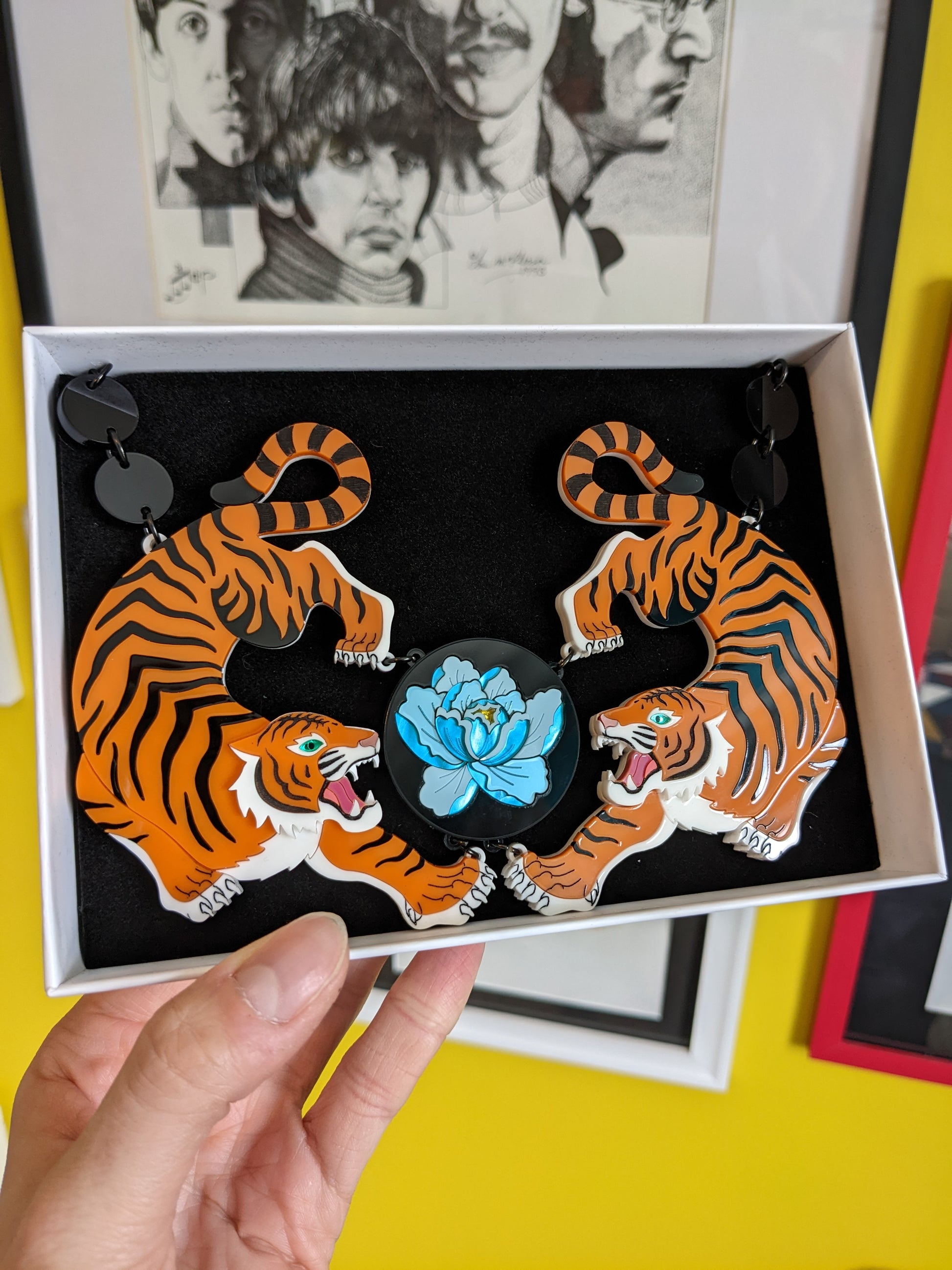 A striking orange and black acrylic necklace featuring two tiger designs with hand-inked details, and a central peony flower motif. The piece is displayed against a pink background with a floral pattern.