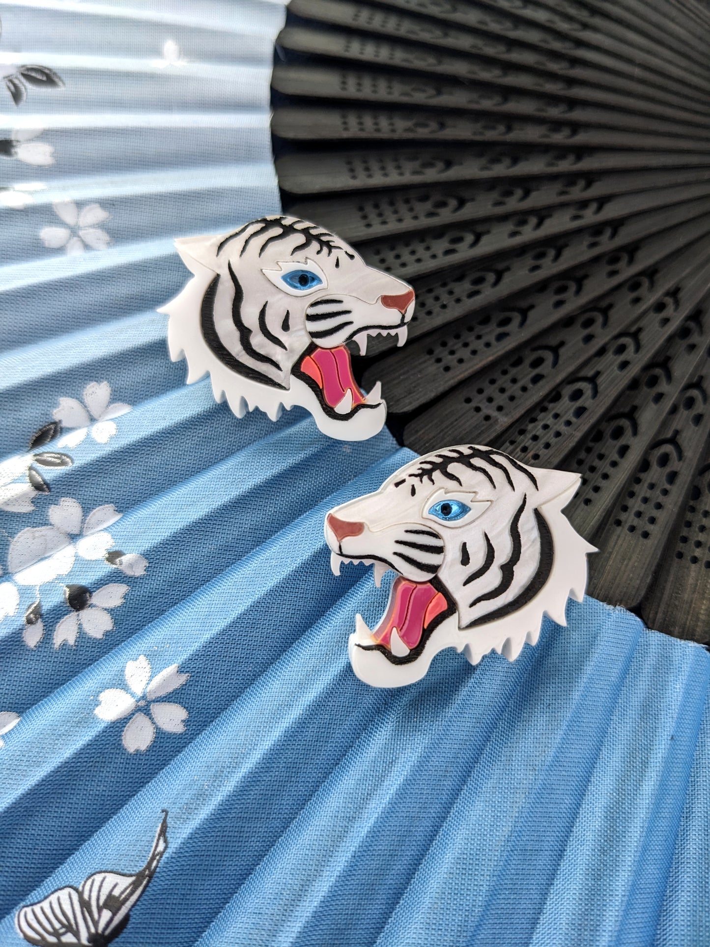 A pair of white tiger stud earrings with blue acrylic eyes and rose gold metallic noses, displayed against a patterned background.