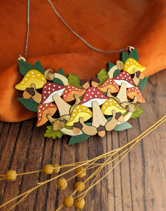 A toadstool-shaped statement bib necklace with autumnal colors, displayed on a wooden surface with a orange cloth in the background.