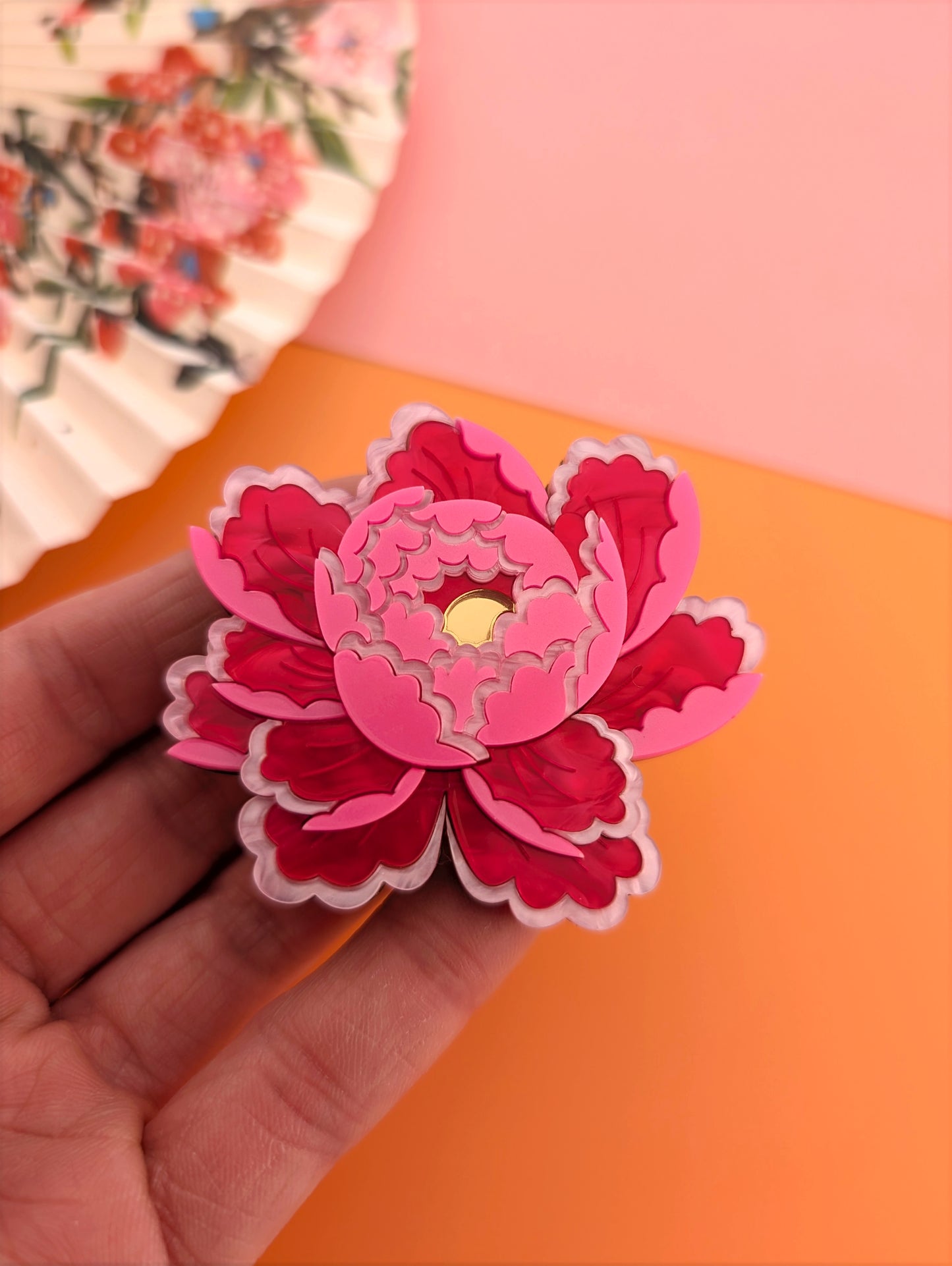 A bright pink peony-inspired flower brooch held in a hand, with layers of petals and a gold mirror centre, against a floral patterned background.
