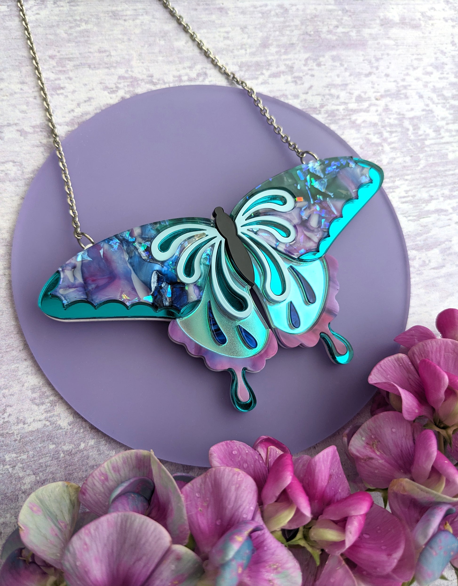 A butterfly necklace with recycled acrylic in purple, teal, and blue, displayed on a chain with a background of purple and pink flowers.