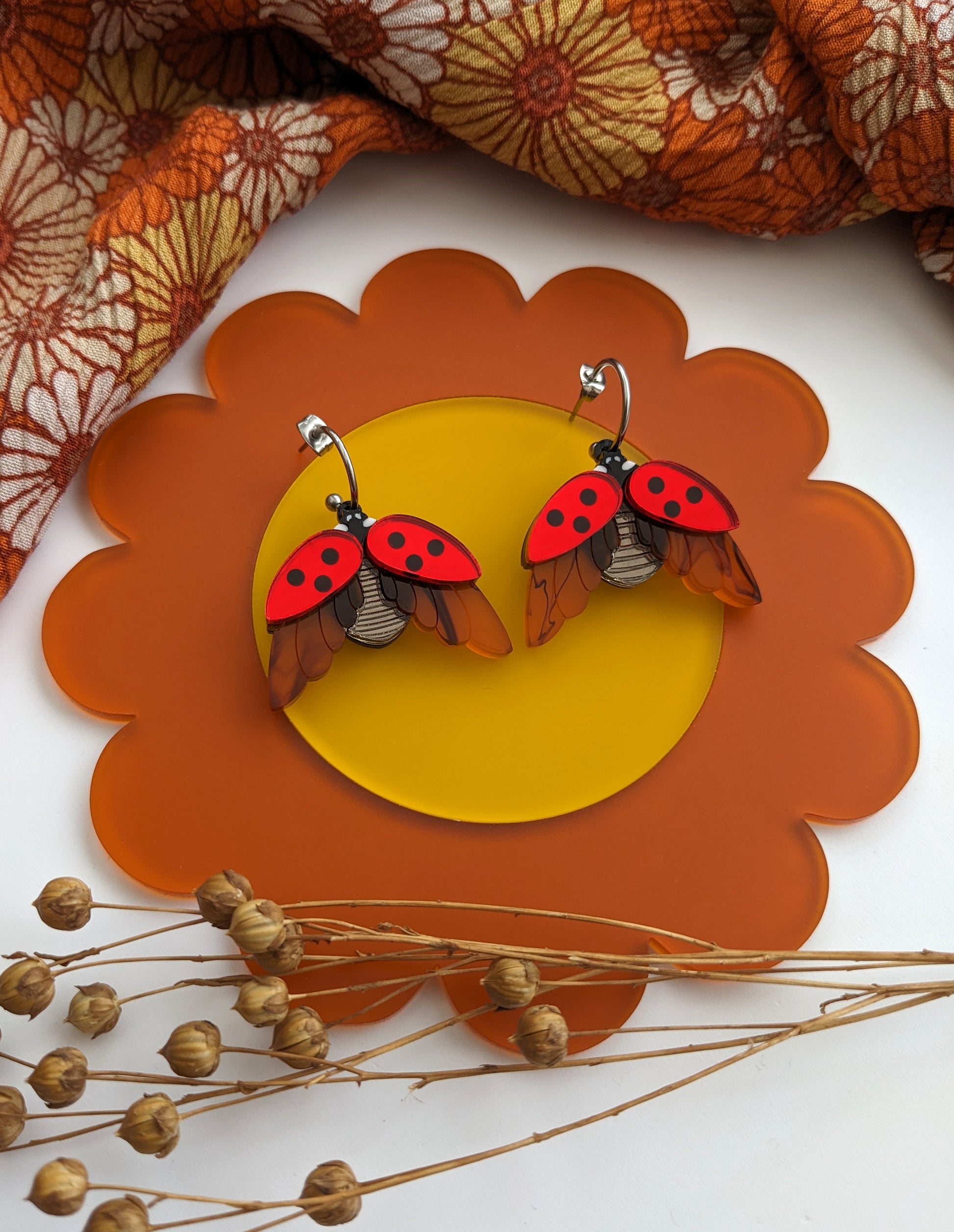 A pair of red and black acrylic ladybird earrings with mirror finish, displayed on a orange and yellow floral patterned surface alongside some decorative elements.