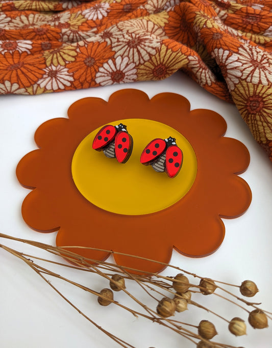 A pair of red ladybird acrylic stud earrings displayed on a yellow and orange circular base, resembling a flower, with a patterned fabric in the background.