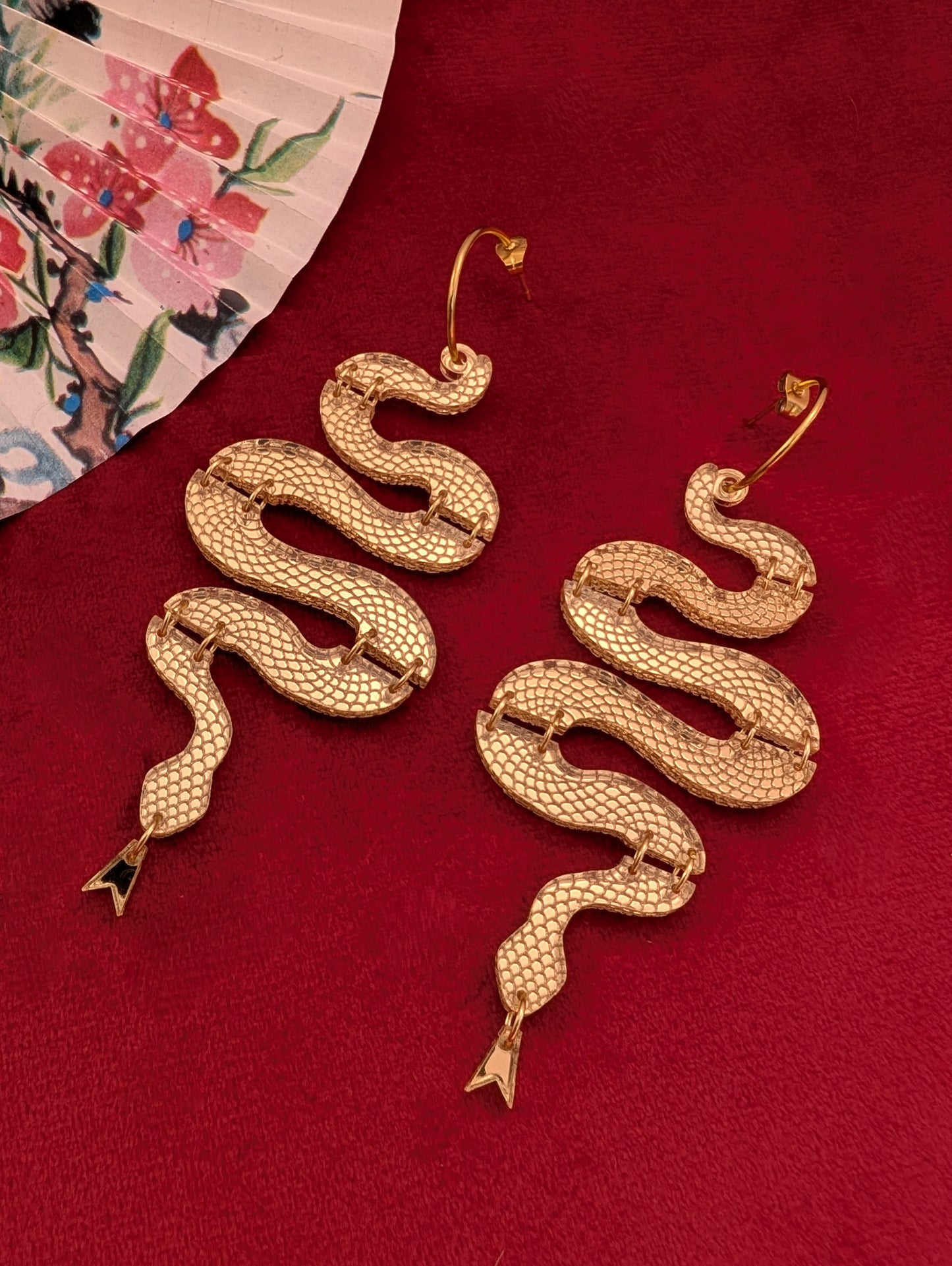 A pair of gold-tone statement snake earrings displayed on a red surface, with one earring slightly above and to the side of the other, near a floral-patterned object at the top left corner.