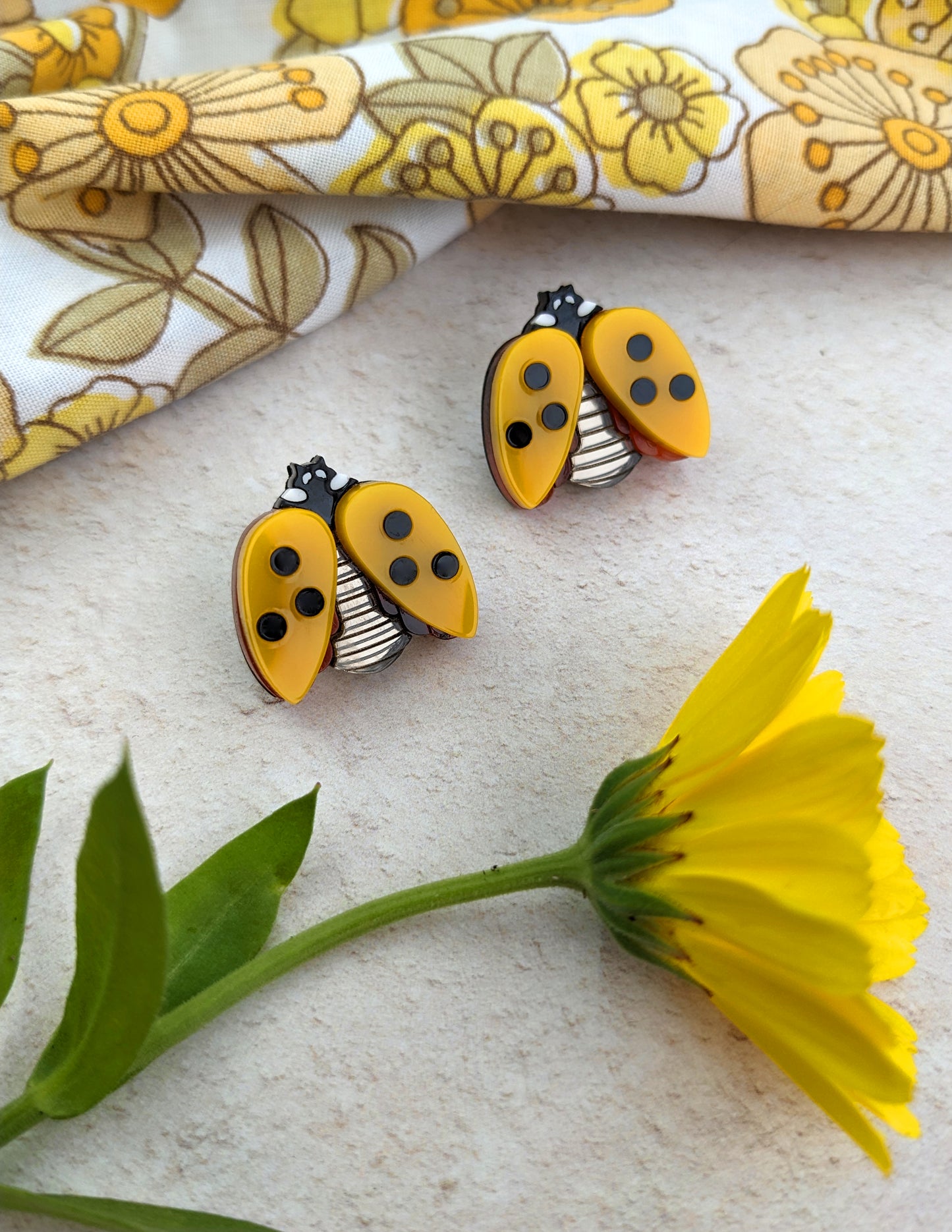 A pair of yellow acrylic ladybird stud earrings with black spots, displayed next to a yellow flower on a patterned fabric.