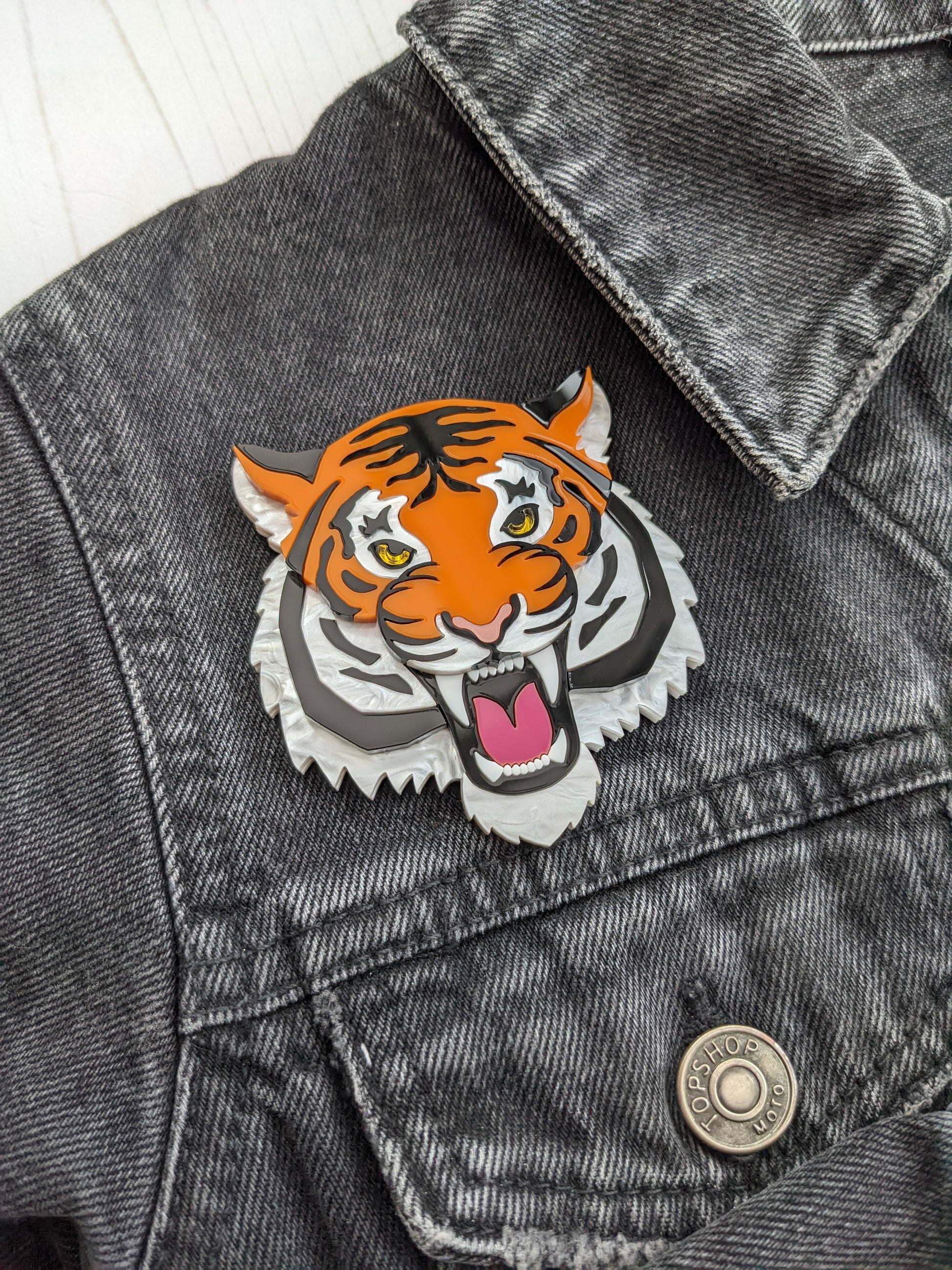 A fierce tiger brooch in orange and white colors pinned onto a gray denim jacket.