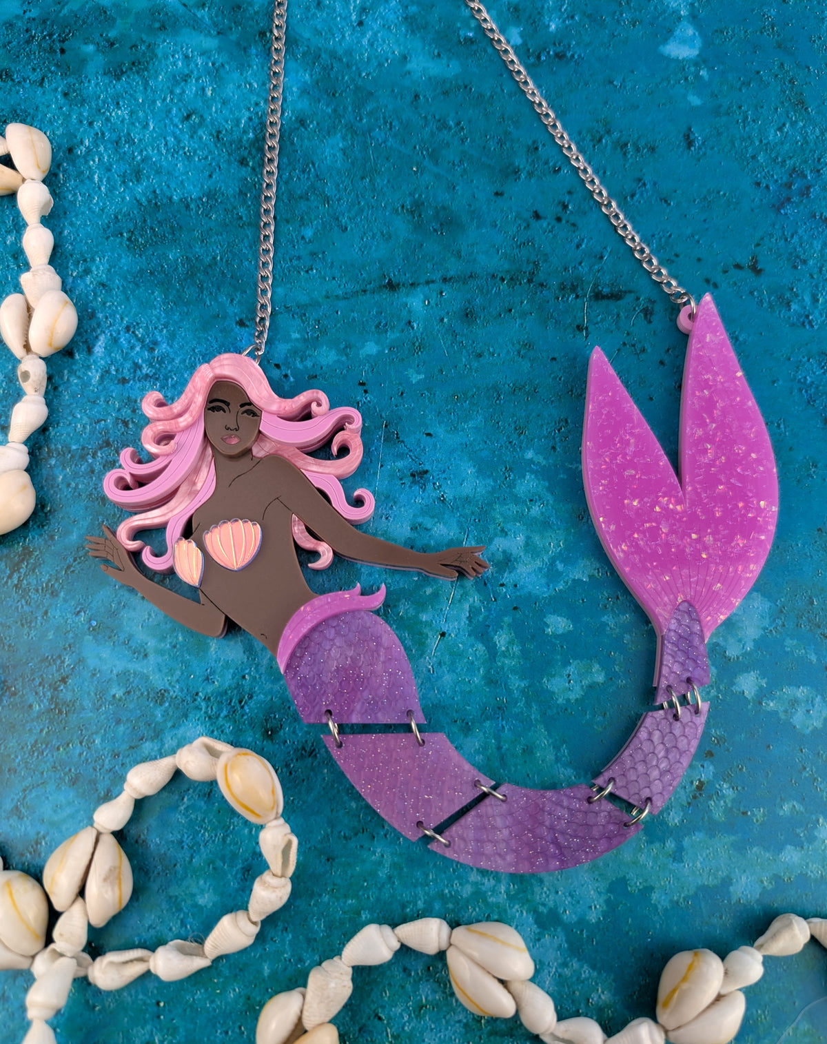 An acrylic mermaid necklace with a pink-haired mermaid figure and a glittery purple tail, displayed on a textured blue background with decorative seashells.