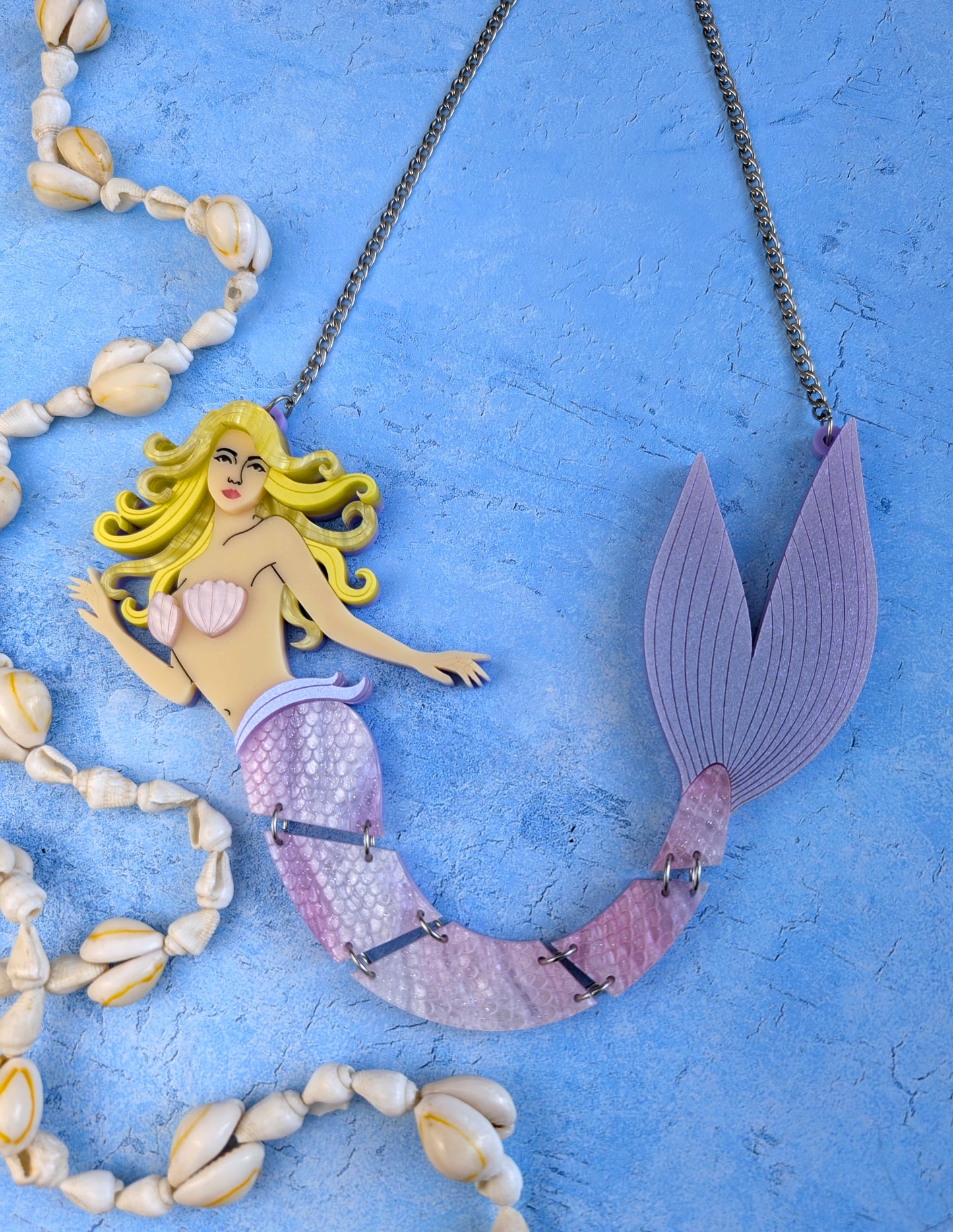A mermaid-themed necklace featuring a yellow-haired mermaid figure with a pink and purple scaled tail, displayed on a textured blue background with decorative seashells.