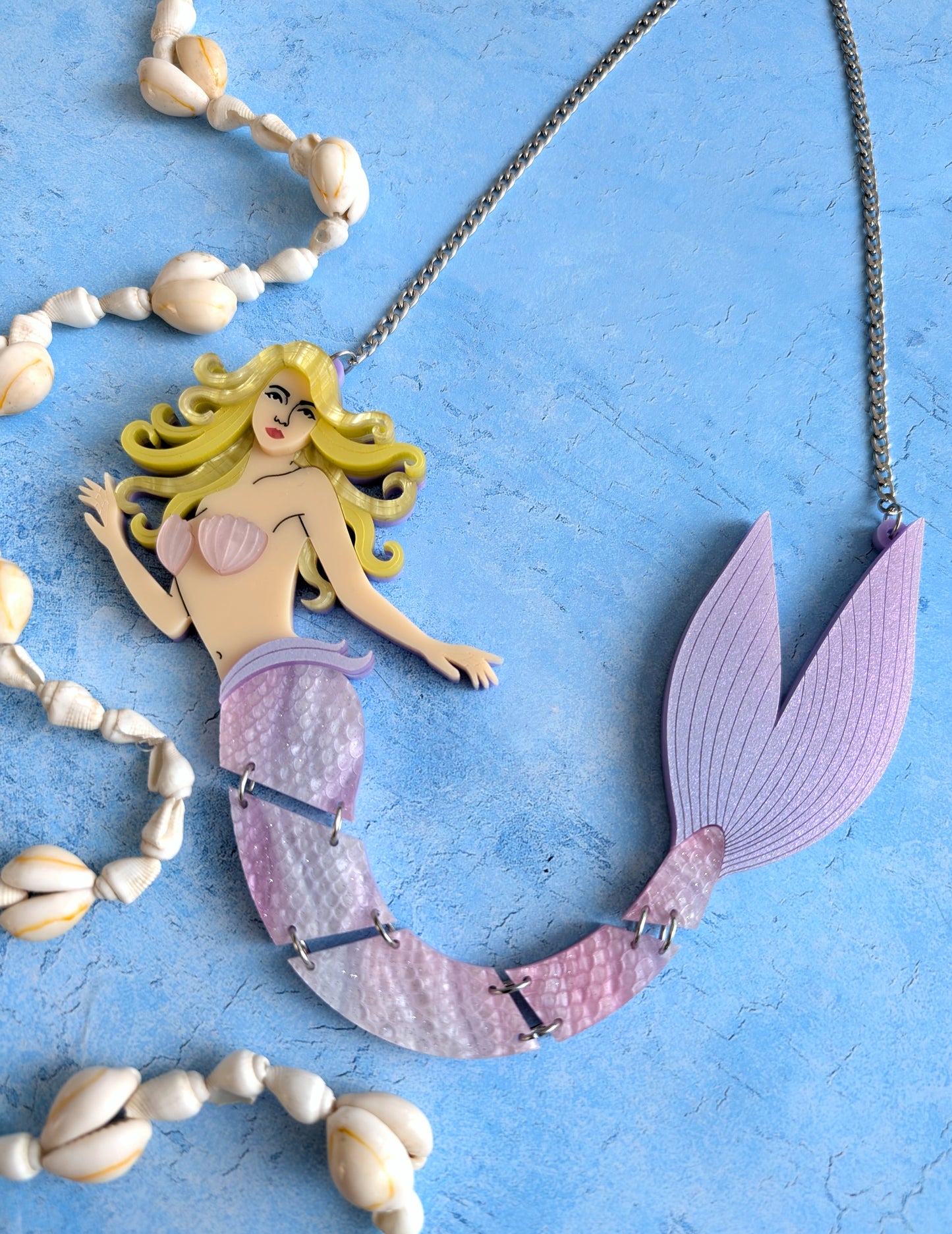 An acrylic mermaid necklace featuring a detailed mermaid figure with yellow hair, a pink shell top, and a purple tail, displayed on a textured blue background with decorative seashells.