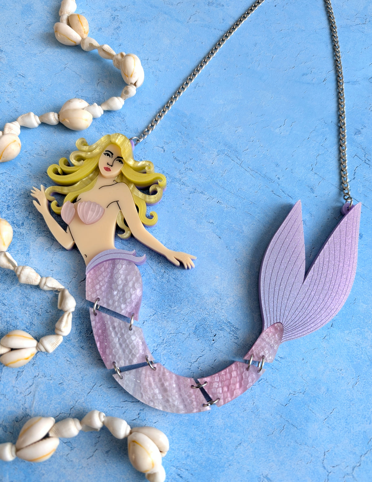 An acrylic mermaid necklace featuring a detailed mermaid figure with yellow hair, a pink shell top, and a purple tail, displayed on a textured blue background with decorative seashells.