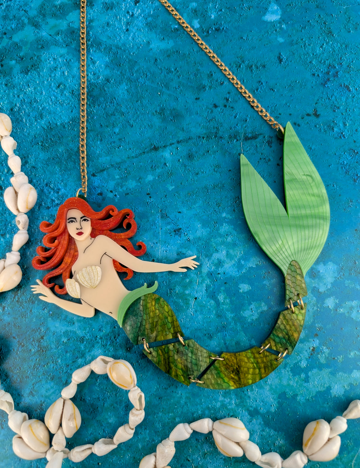 A laser cut handmade mermaid acrylic necklace featuring a mermaid with copper red hair, a shell top, and a green scaled tail, hanging from a gold-tone chain against a textured blue background with seashell garlands.