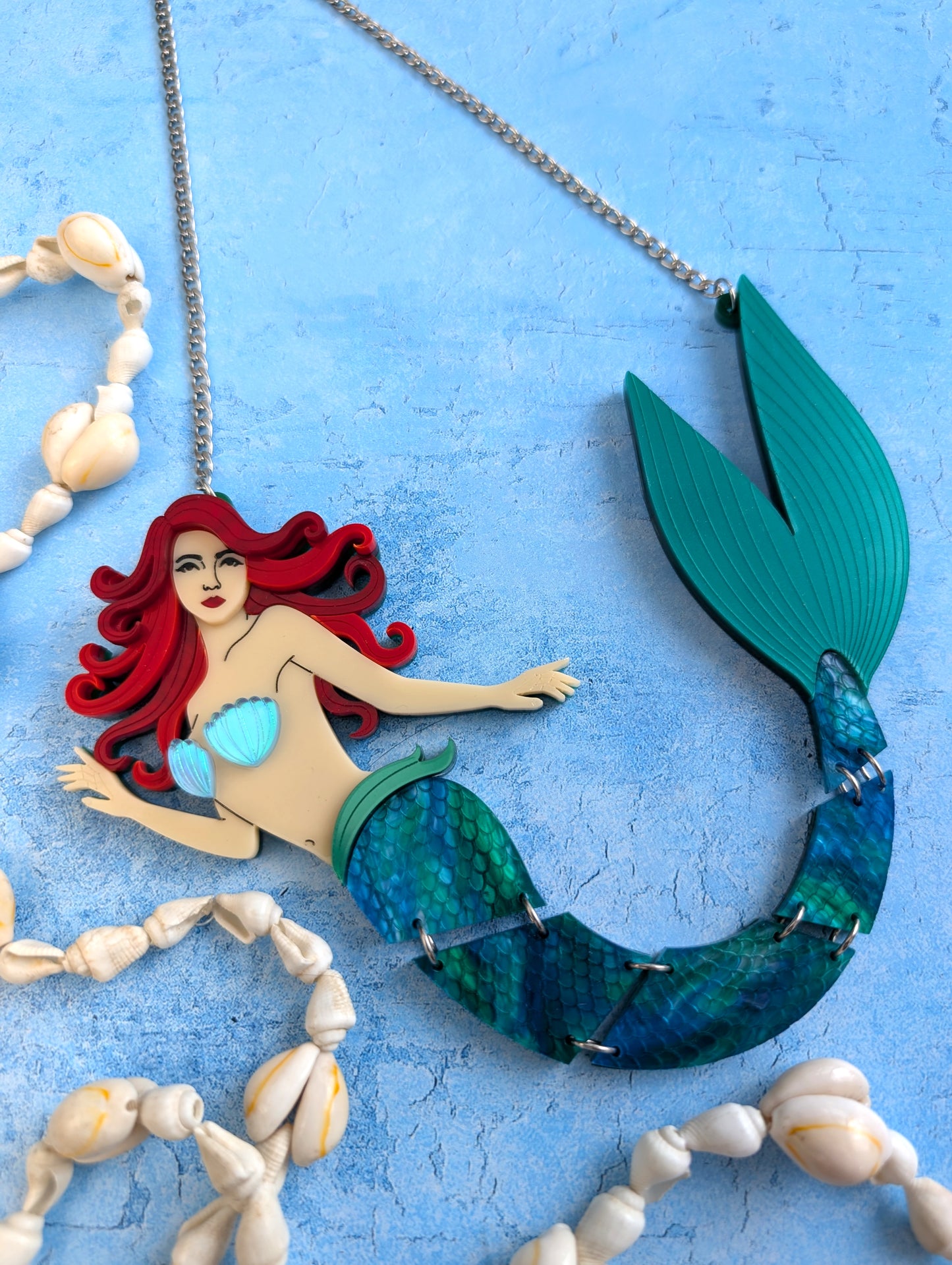 An acrylic mermaid necklace featuring a red-haired mermaid with a teal and blue scaled tail, displayed on a textured light blue background with decorative seashells.