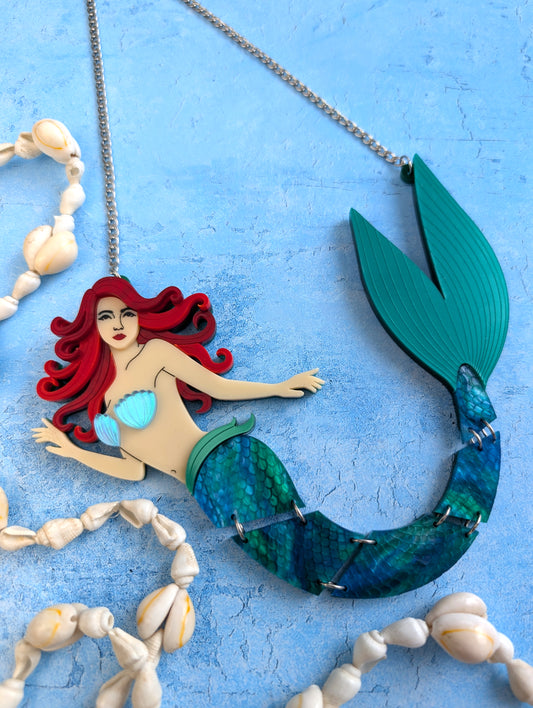An acrylic mermaid necklace featuring a red-haired mermaid with a teal and blue scaled tail, displayed on a textured light blue background with decorative seashells.