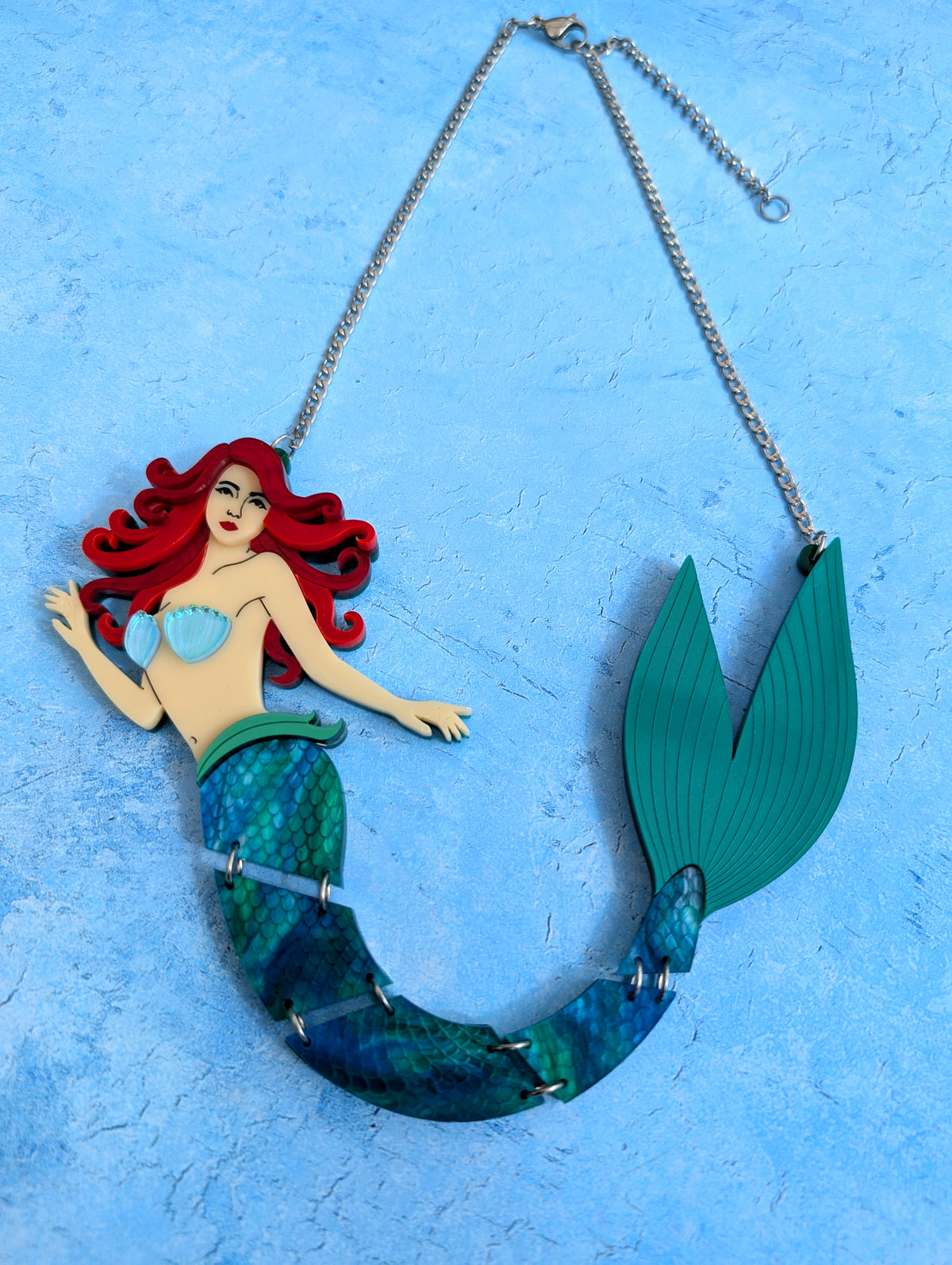 A mermaid-themed acrylic necklace featuring a red-haired mermaid with a teal and blue scaled tail, suspended from a stainless steel chain against a textured light blue background.