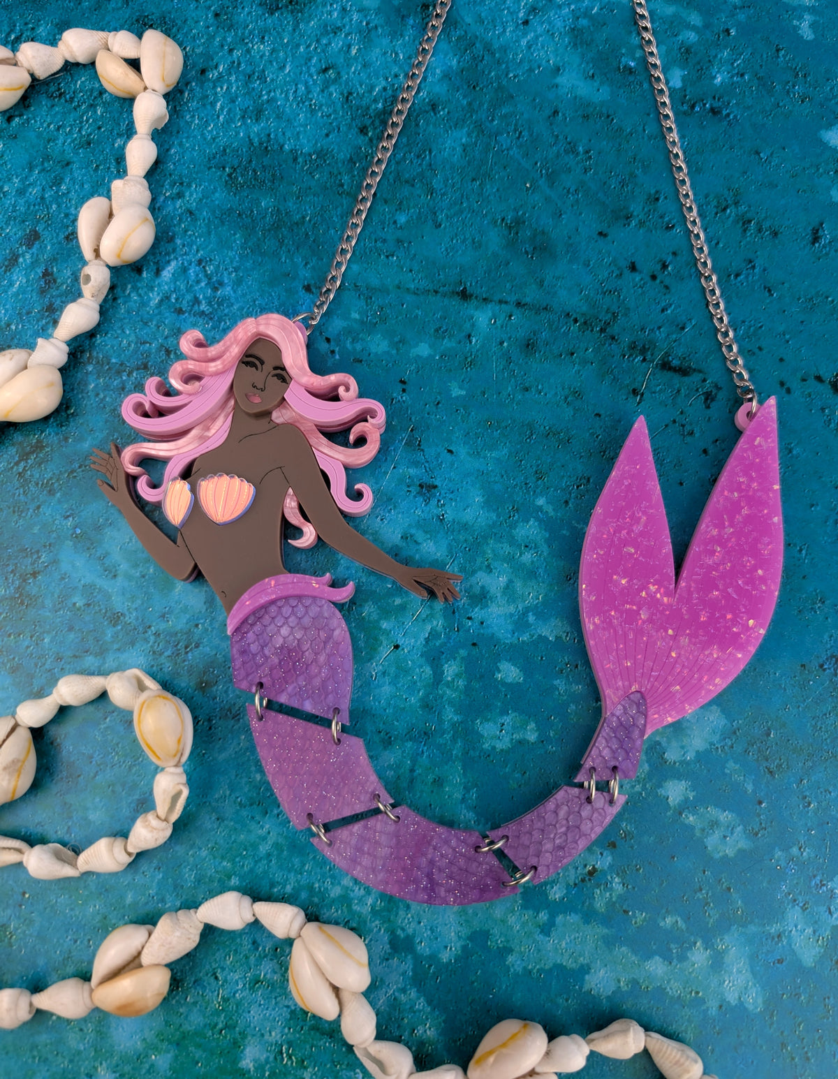 An acrylic mermaid necklace featuring a stylized mermaid figure with pink hair, a pink shell top, and a purple-to-pink gradient tail, hanging from a silver chain on a textured blue background with seashell garlands.