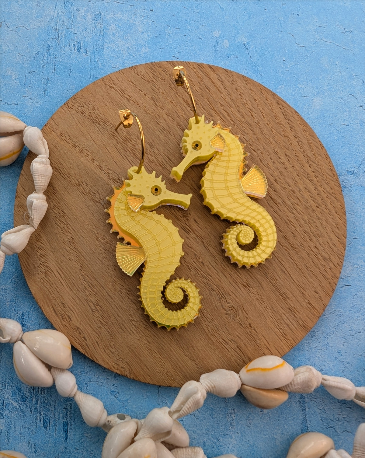 A pair of yellow seahorse-shaped earrings displayed on a round wooden board, surrounded by white seashells on a textured blue background.