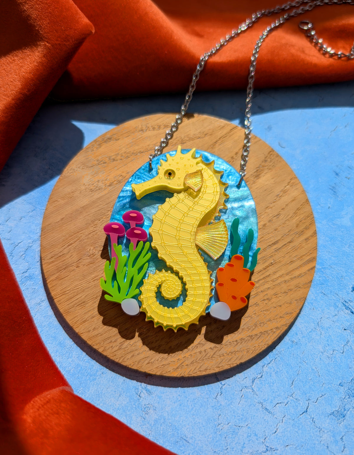 A colourful seahorse pendant necklace displayed on a round wooden board. The pendant features a bright yellow seahorse on a blue underwater-themed background with small green, pink, and orange sea plants.