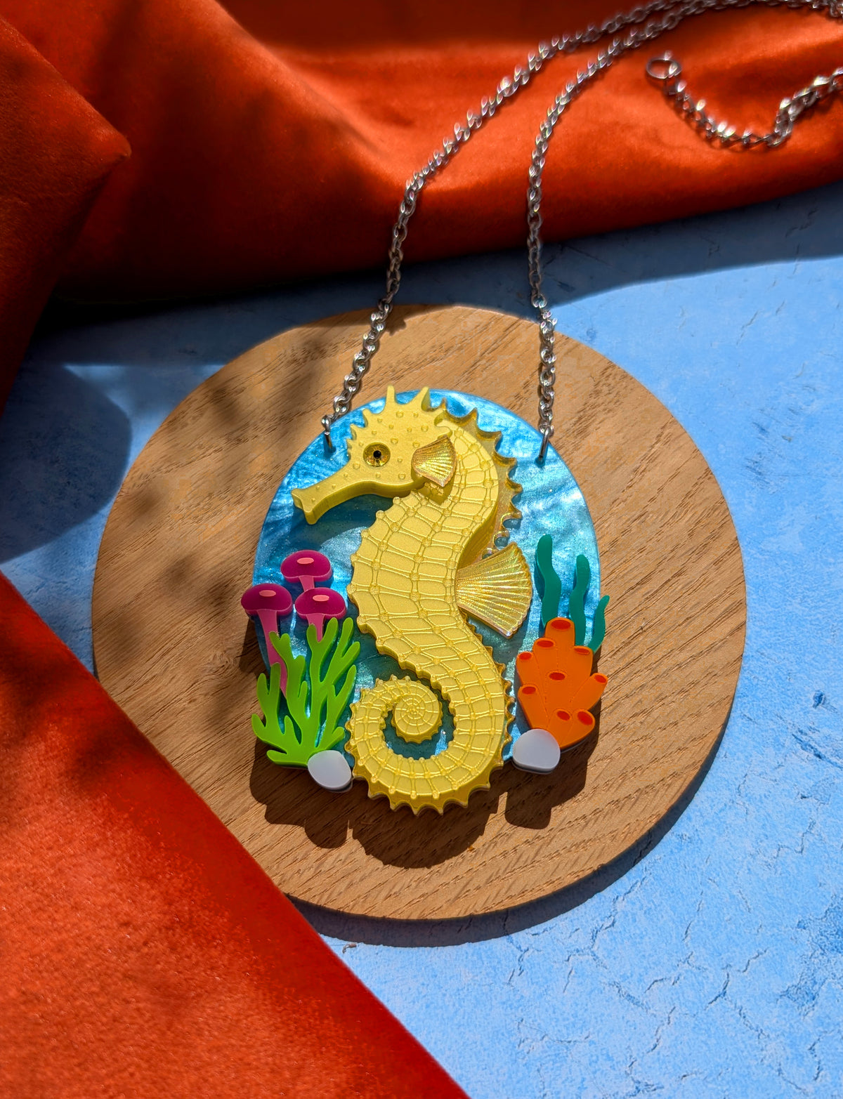 A colourful seahorse pendant necklace displayed on a round wooden board. The pendant features a bright yellow seahorse on a blue underwater-themed background with small green, pink, and orange sea plants.