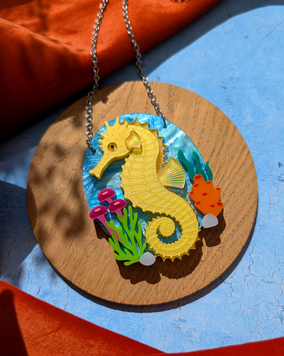 A colourful seahorse pendant necklace displayed on a round wooden board. The pendant features a bright yellow seahorse on a blue underwater-themed background with small green, pink, and orange sea plants.