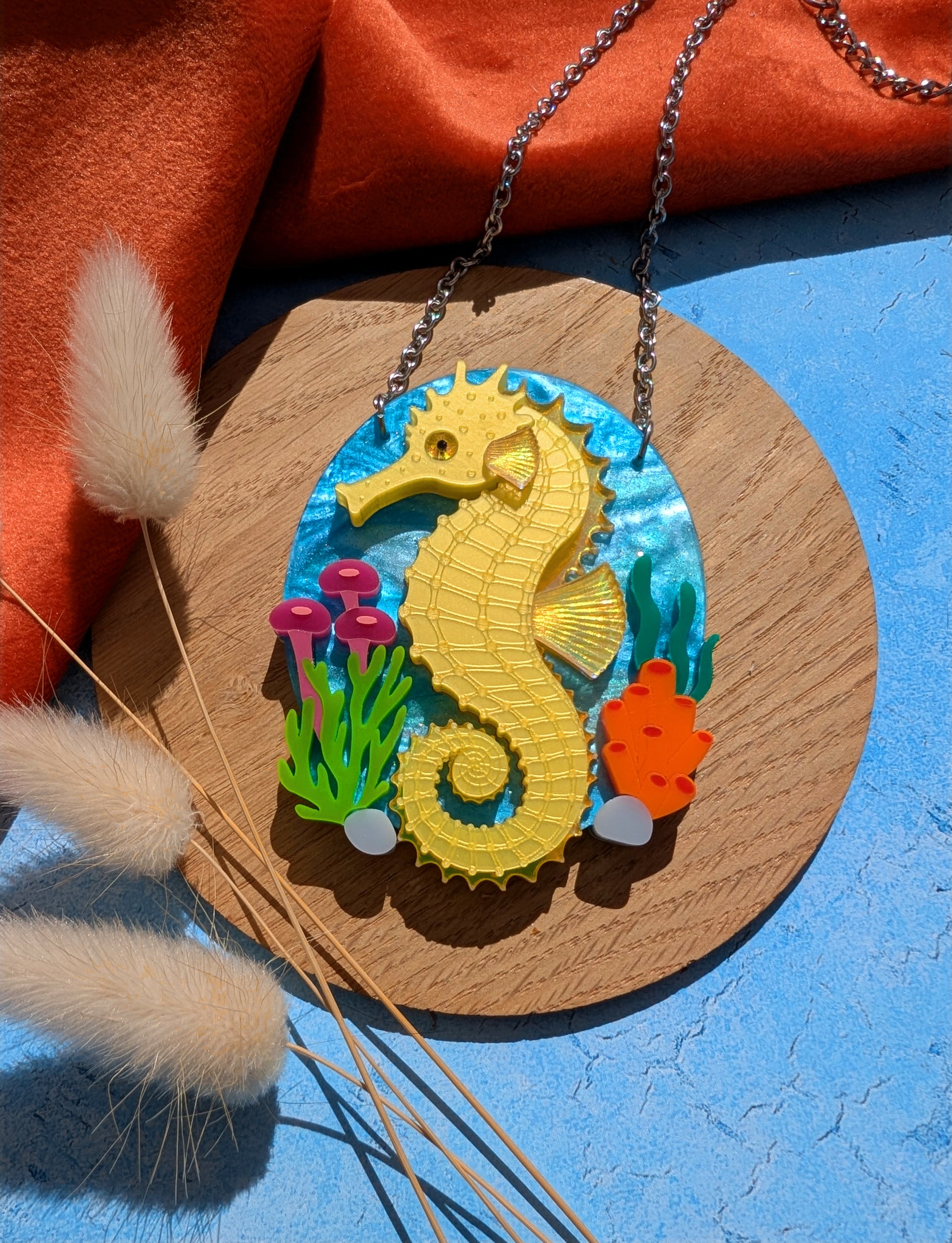 A colourful seahorse pendant necklace displayed on a round wooden board. The pendant features a bright yellow seahorse on a blue underwater-themed background with small green, pink, and orange sea plants.
