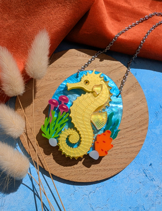 A colourful seahorse pendant necklace displayed on a round wooden board. The pendant features a bright yellow seahorse on a blue underwater-themed background with small green, pink, and orange sea plants.